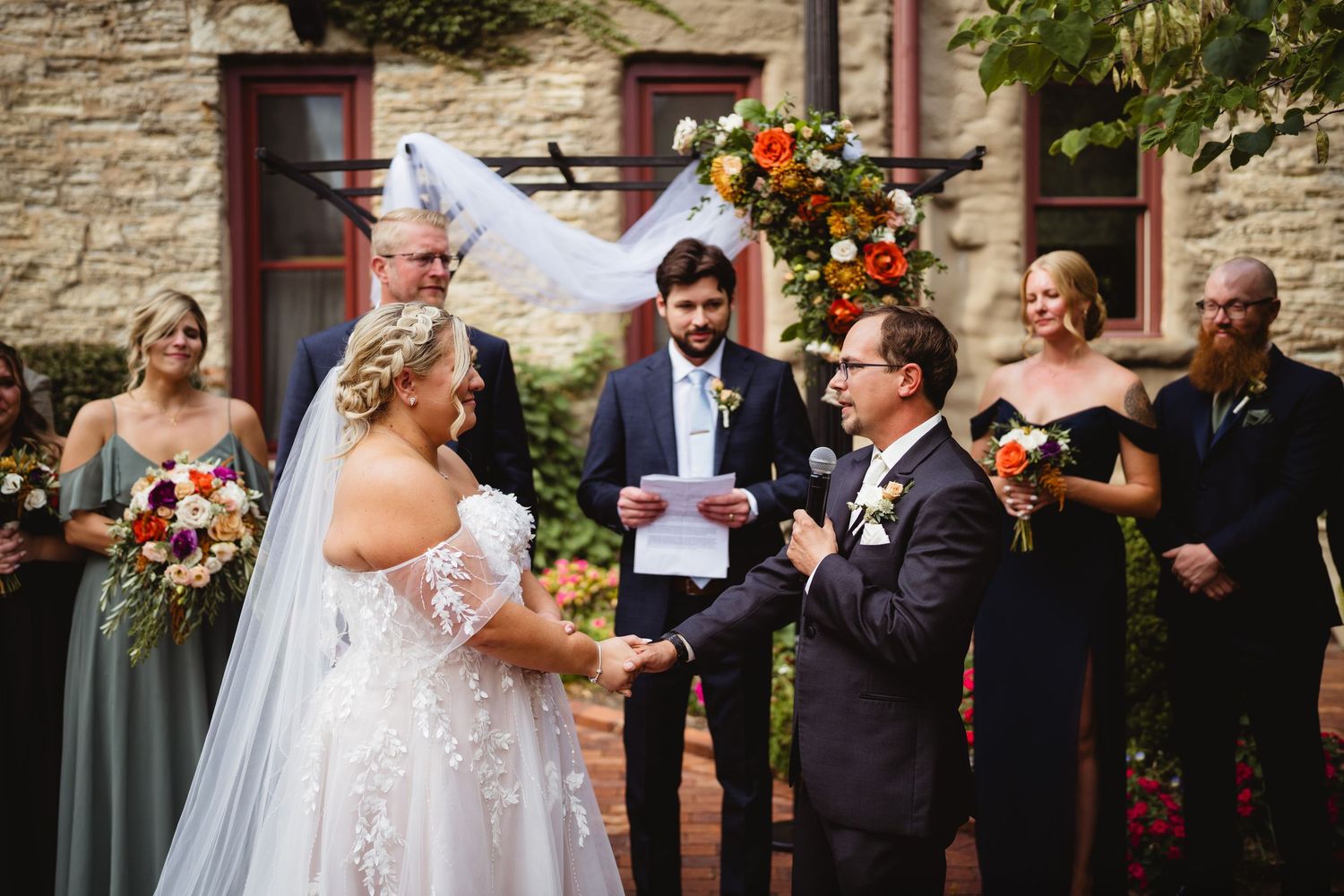 A romantic outdoor wedding ceremony moment takes place in front of a rustic stone wall adorned with flowers.