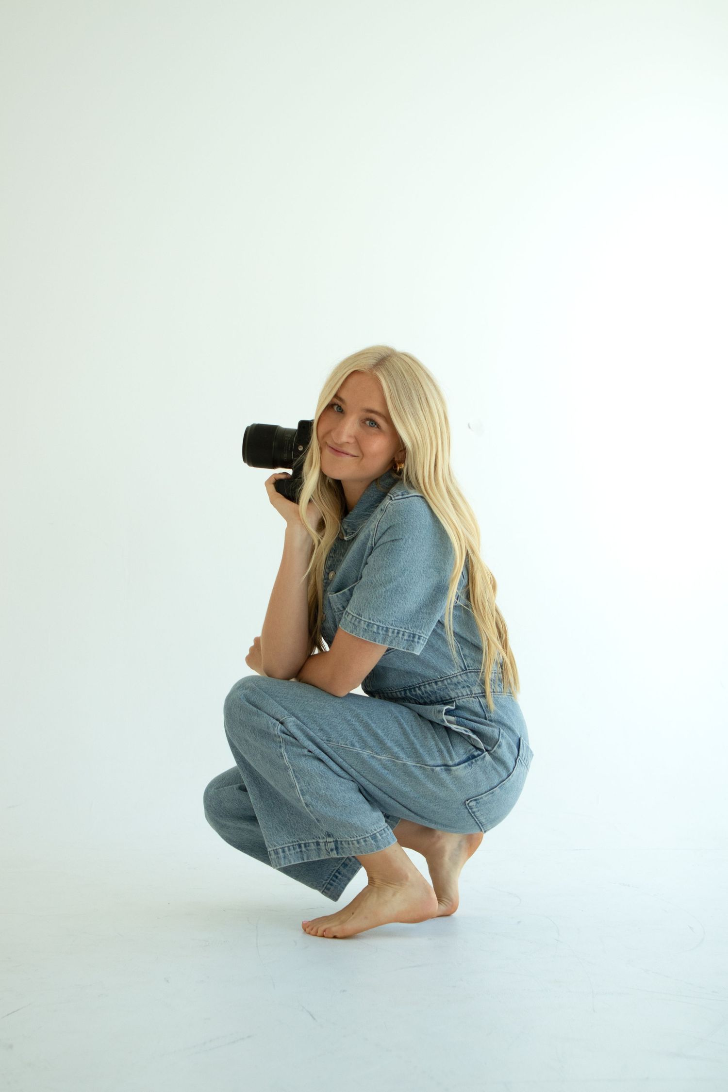 Photographer in denim outfit crouches with camera against white background.