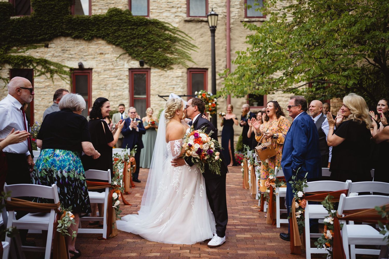 Wedding party stands in a row against stone wall with greenery during outdoor ceremony at rustic venue.