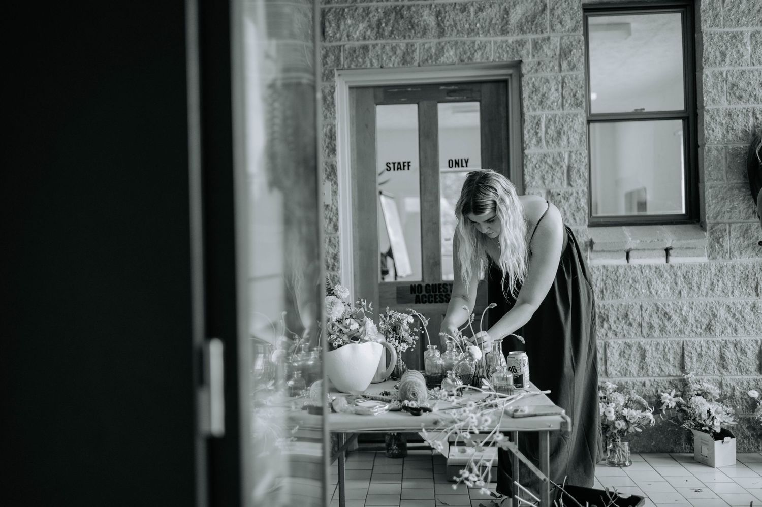 A person arranges plants and items on a table outside a stone building in this vintage black and white photograph.