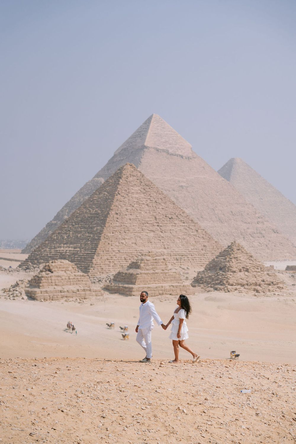 A couple in white outfits walking hand in hand across the desert landscape with the pyramids of Giza visible behind them.