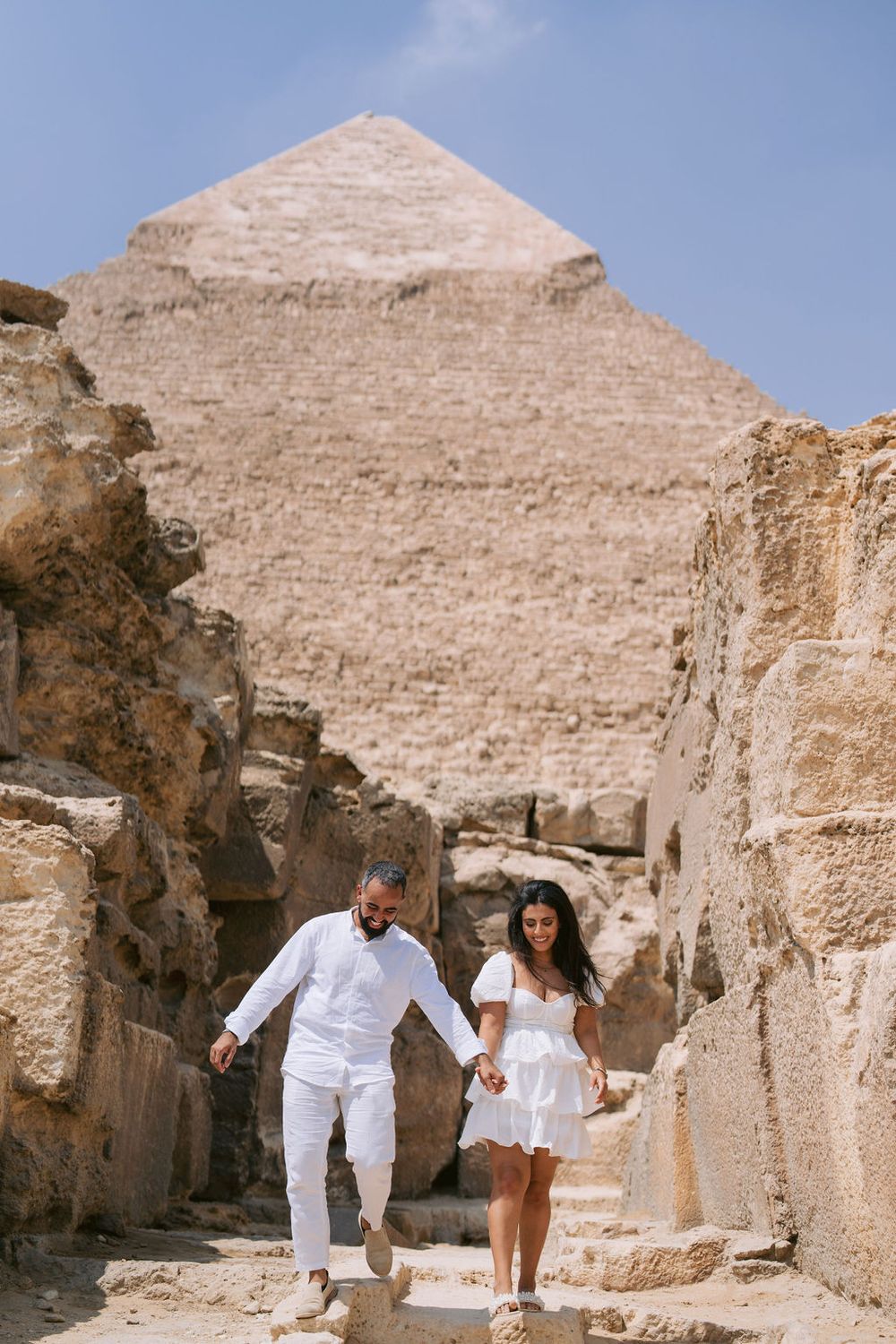 Couple in white outfits exploring ancient stone ruins with pyramids visible in the desert background.