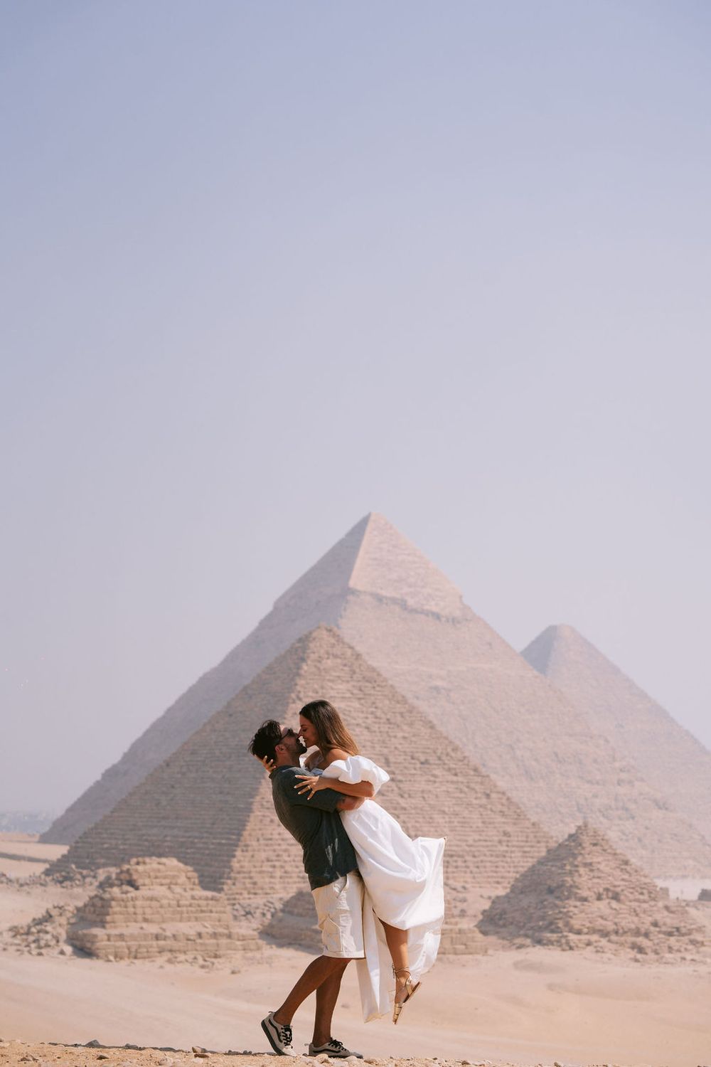 A romantic couple dances and embraces in front of the ancient Egyptian pyramids of Giza on a hazy desert day.