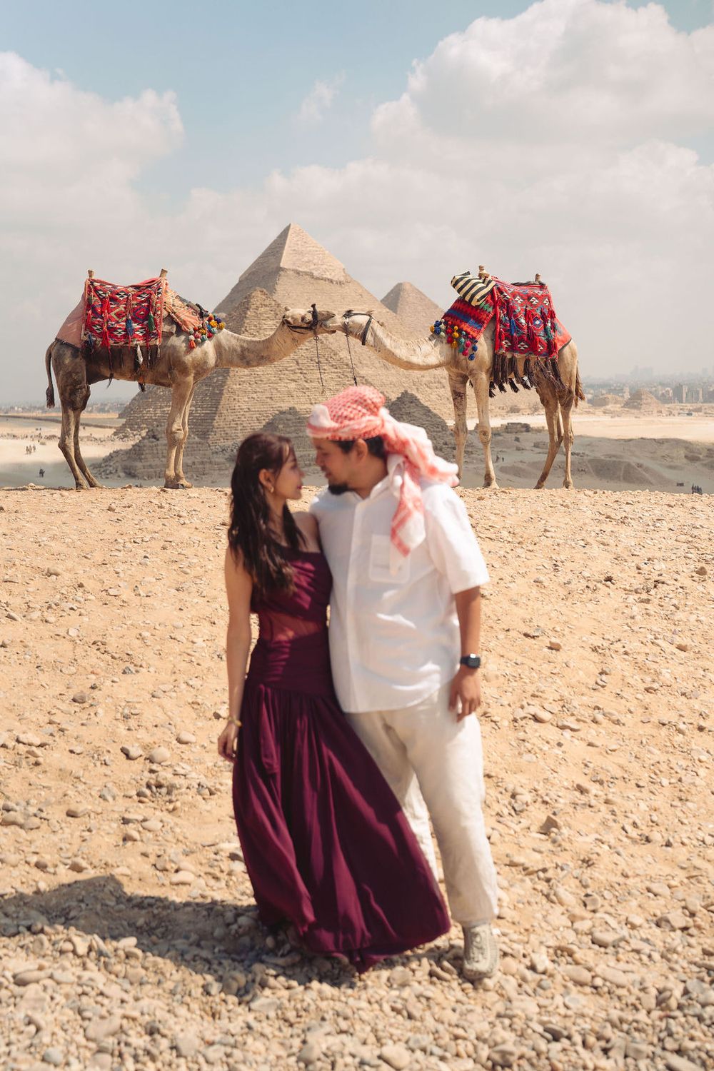 A couple embraces in front of the Great Pyramids of Giza while two decorated camels rest in the background.