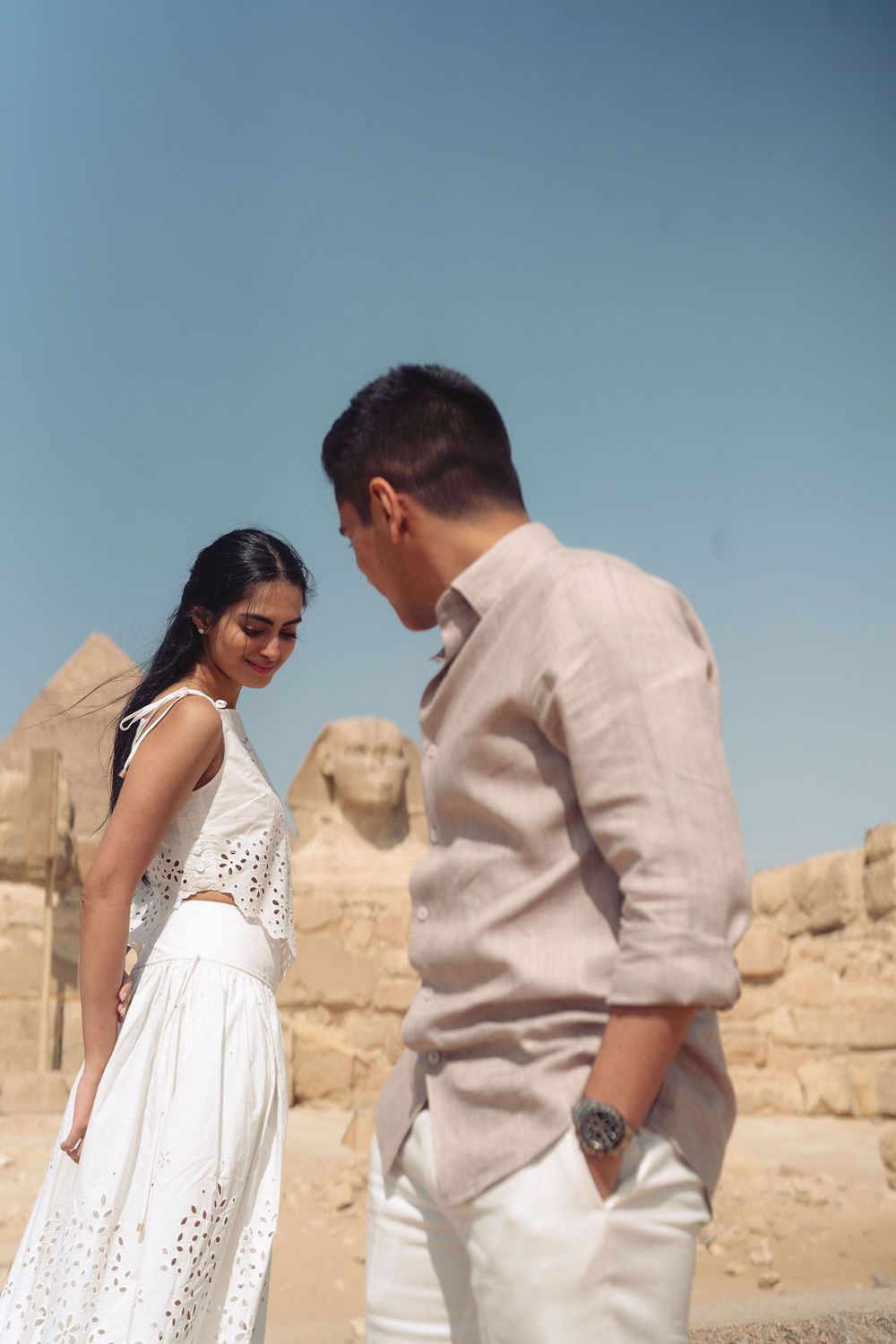 A couple in white and beige outfits share romantic moments against a desert backdrop with ancient ruins.