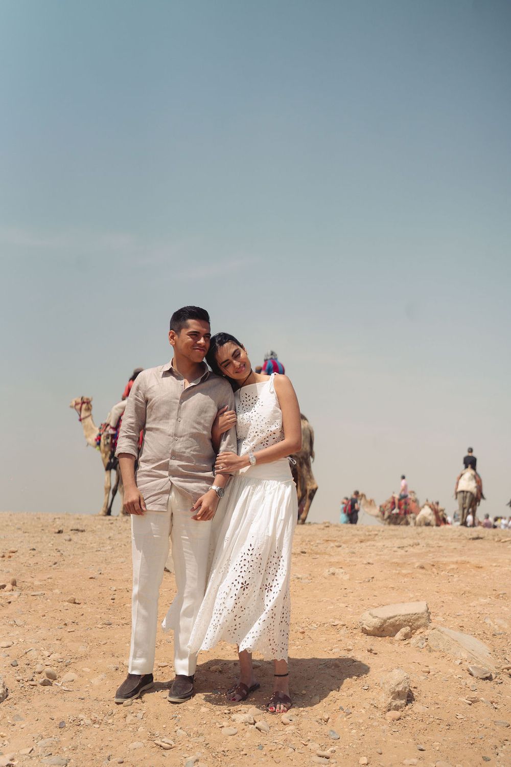 Couple in light-colored outfits posing together on a sandy desert landscape under blue sky.