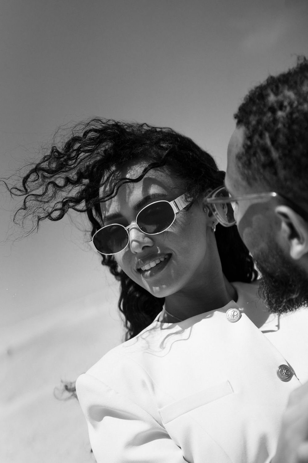 A black and white photo series showing a person with curly hair and sunglasses smiling and laughing against a light background.