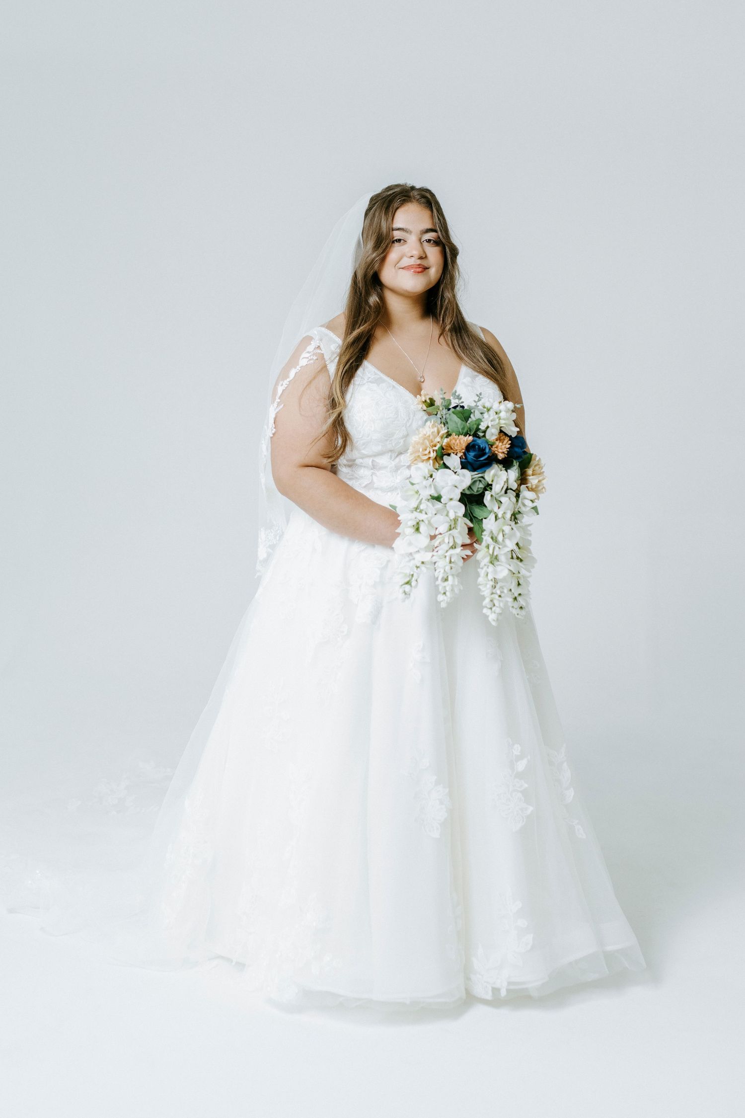 A graceful plus-size bride in a white tulle dress poses against a light backdrop holding delicate floral bouquets.