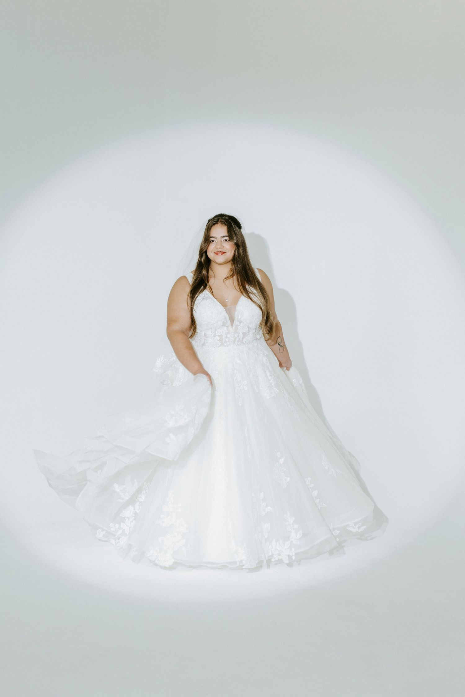 A figure in a flowing white wedding dress poses gracefully against a soft white background in a studio setting.