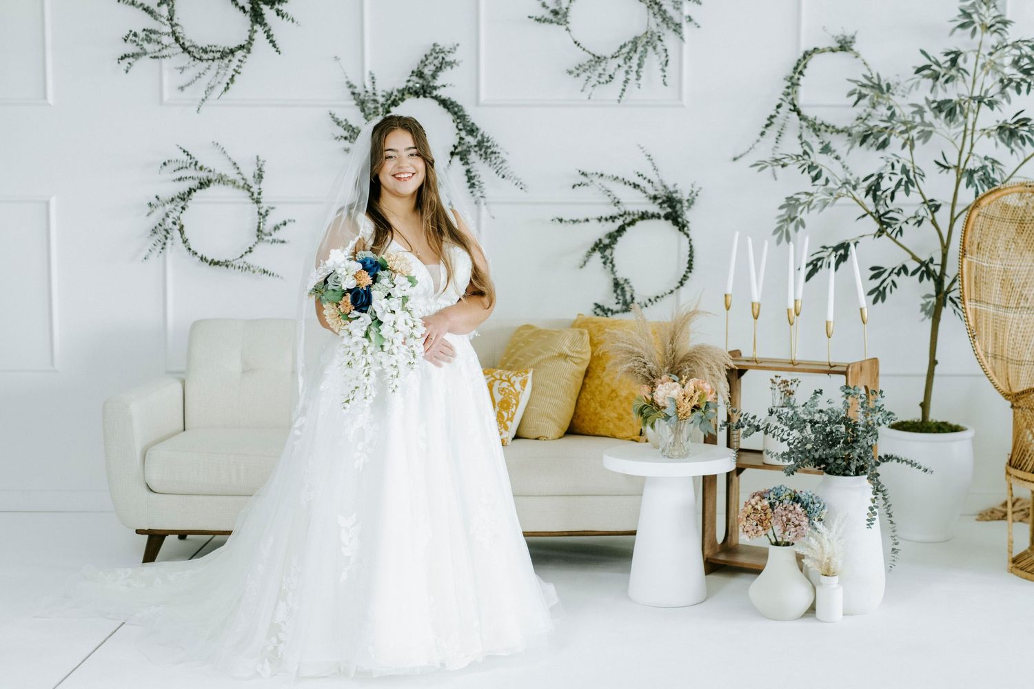 Elegant bride in flowing white gown poses in bright room with floral wallpaper and modern decor.