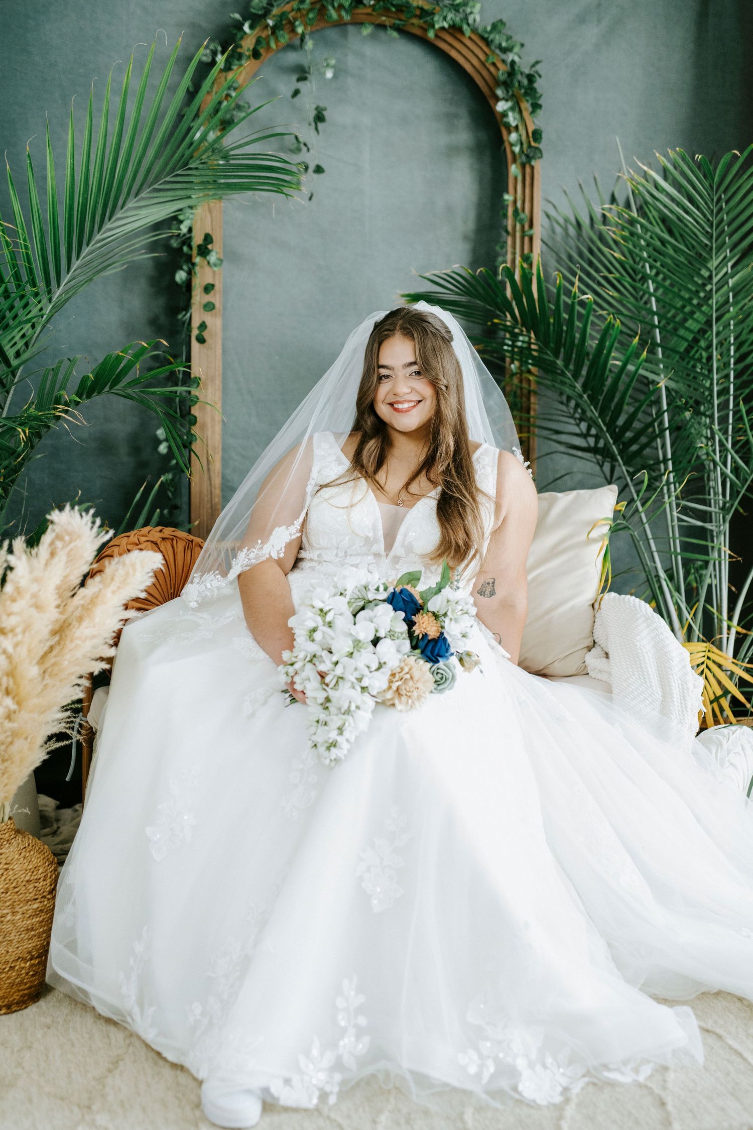 A bridal portrait showcasing a wedding dress and bouquet amid tropical palm fronds and an ornate arch backdrop.