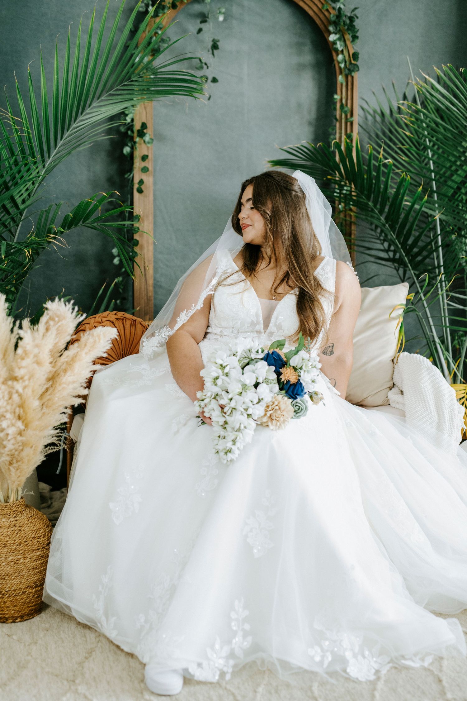 A bride in a flowing white wedding dress poses elegantly on wicker furniture surrounded by tropical palm leaves and pampas grass.