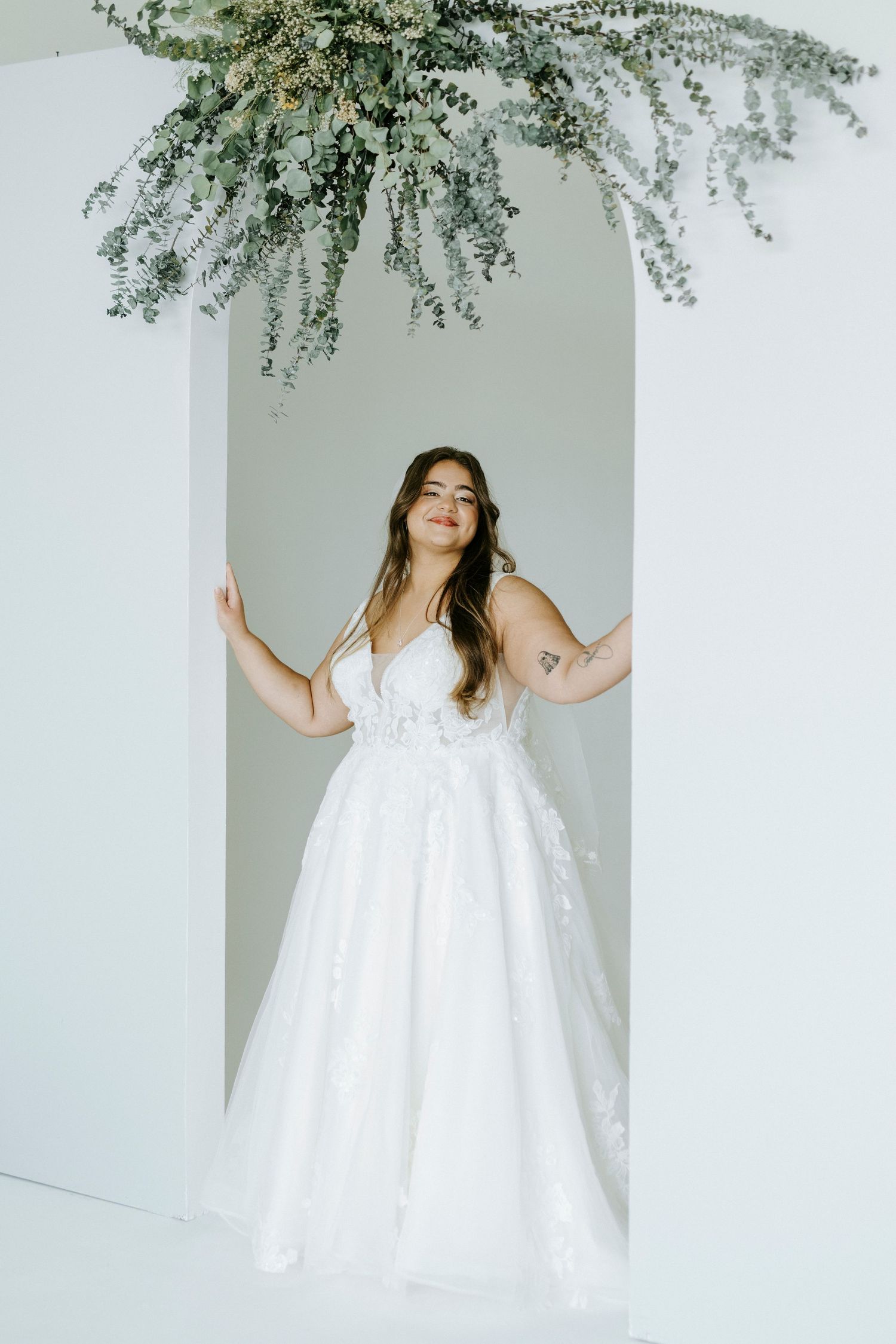 Ethereal bridal photos under hanging greenery with flowing white dress in minimalist studio setting.