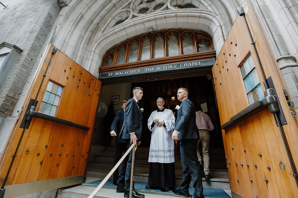 A group stands in the doorway of a grand stone church with wooden doors and gothic architecture.