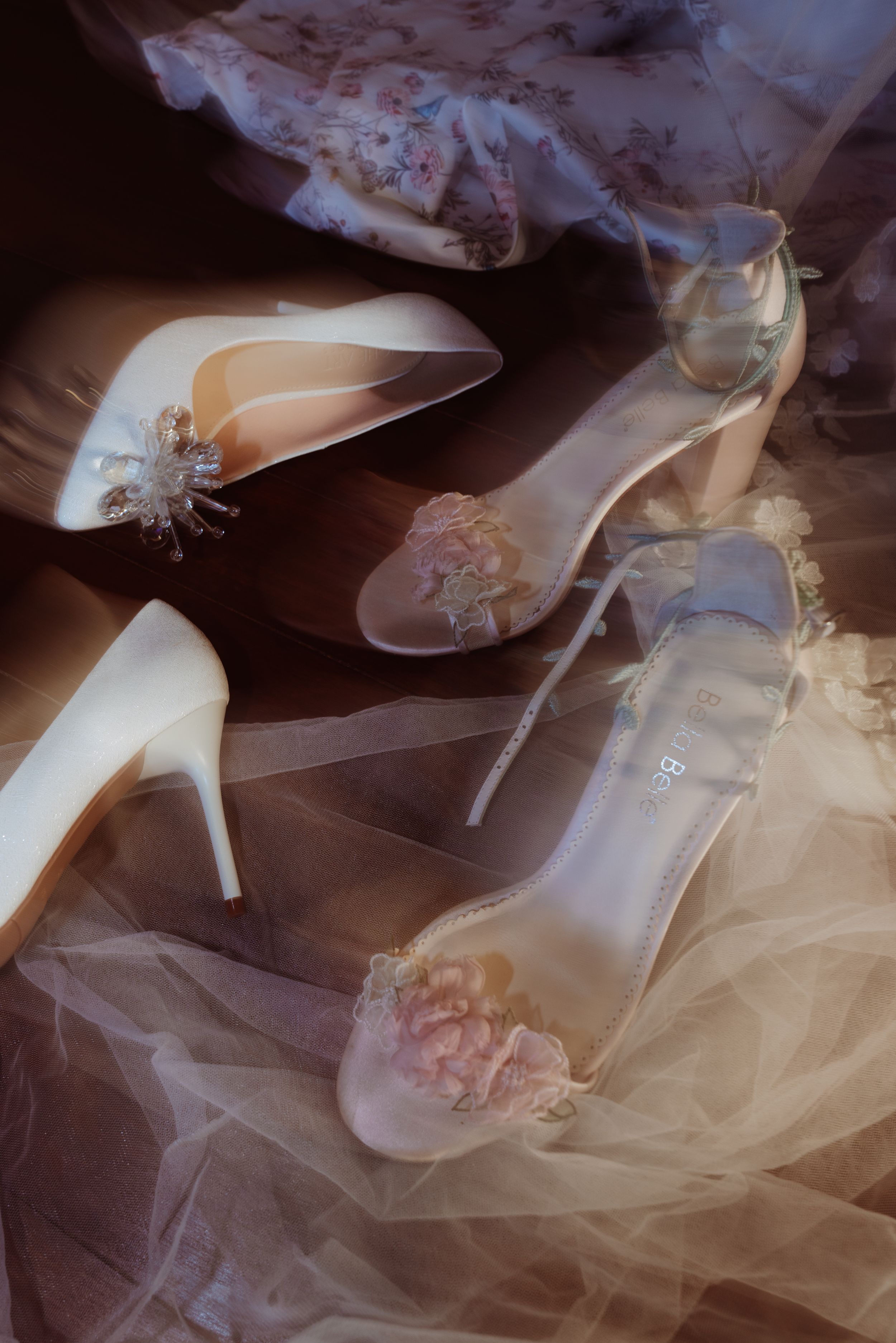 Elegant bridal heels including white pumps and clear floral sandals arranged on soft tulle fabric.