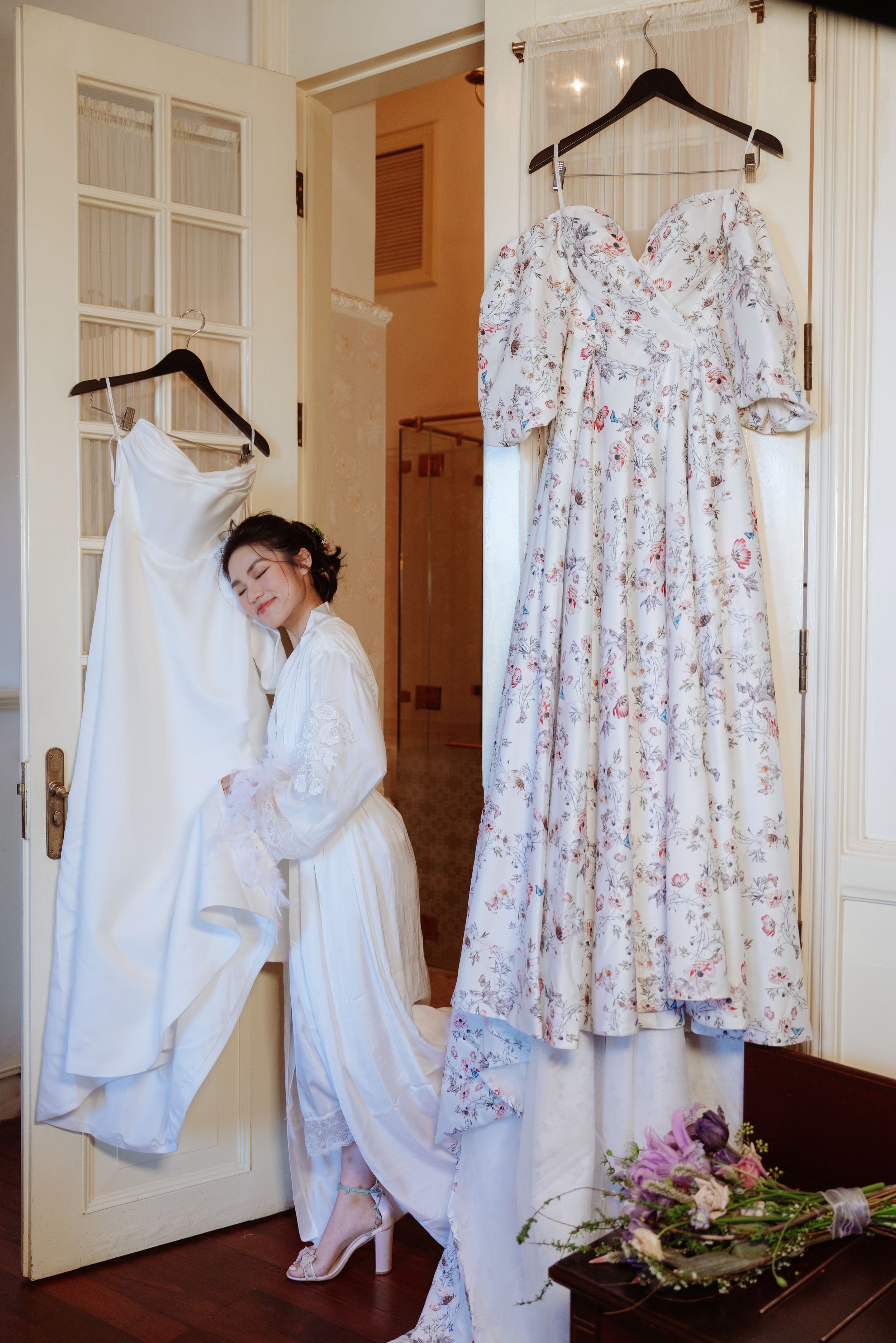 Bride in white robe standing between hanging wedding gown and floral dress in elegant hotel room.