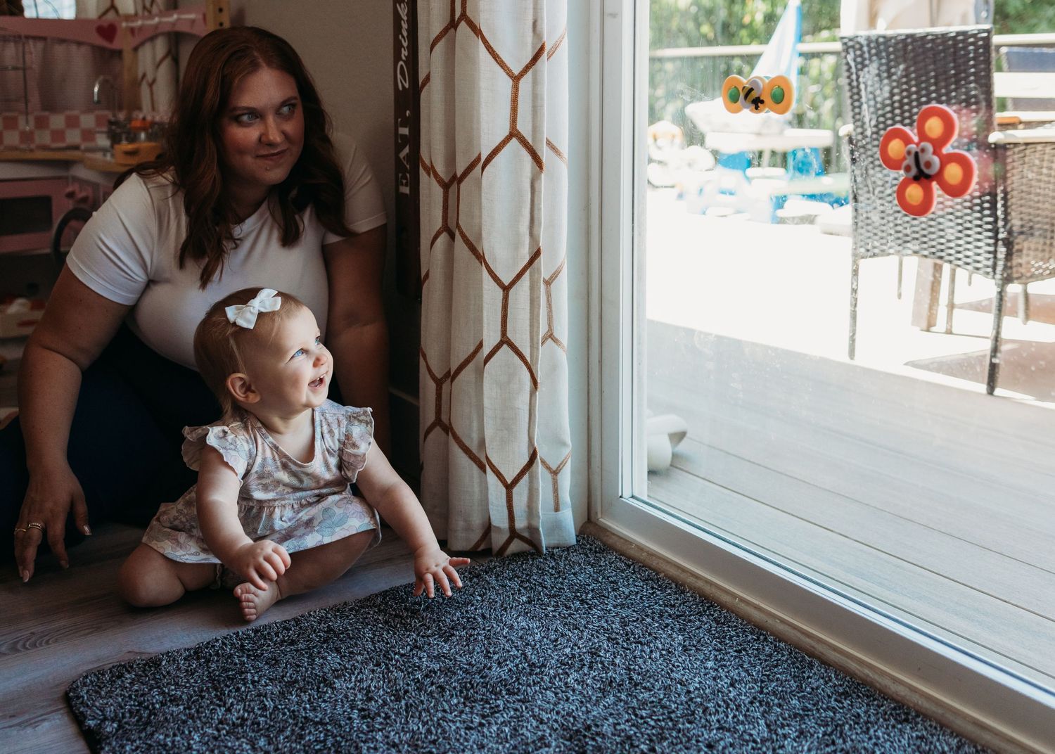 A toddler in a floral dress sits on a gray carpet by a sliding glass door while being supported by someone.