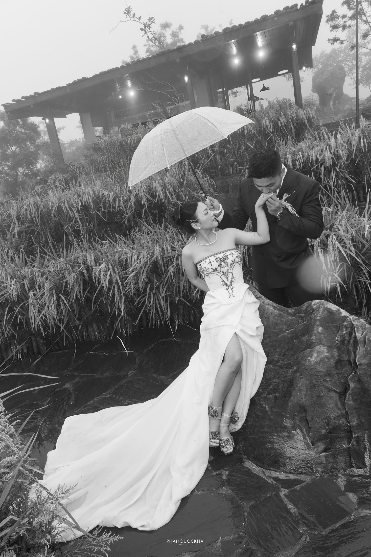 Groom kisses brides hand under umbrella as she laughs on rocks, elegant black and white portrait.