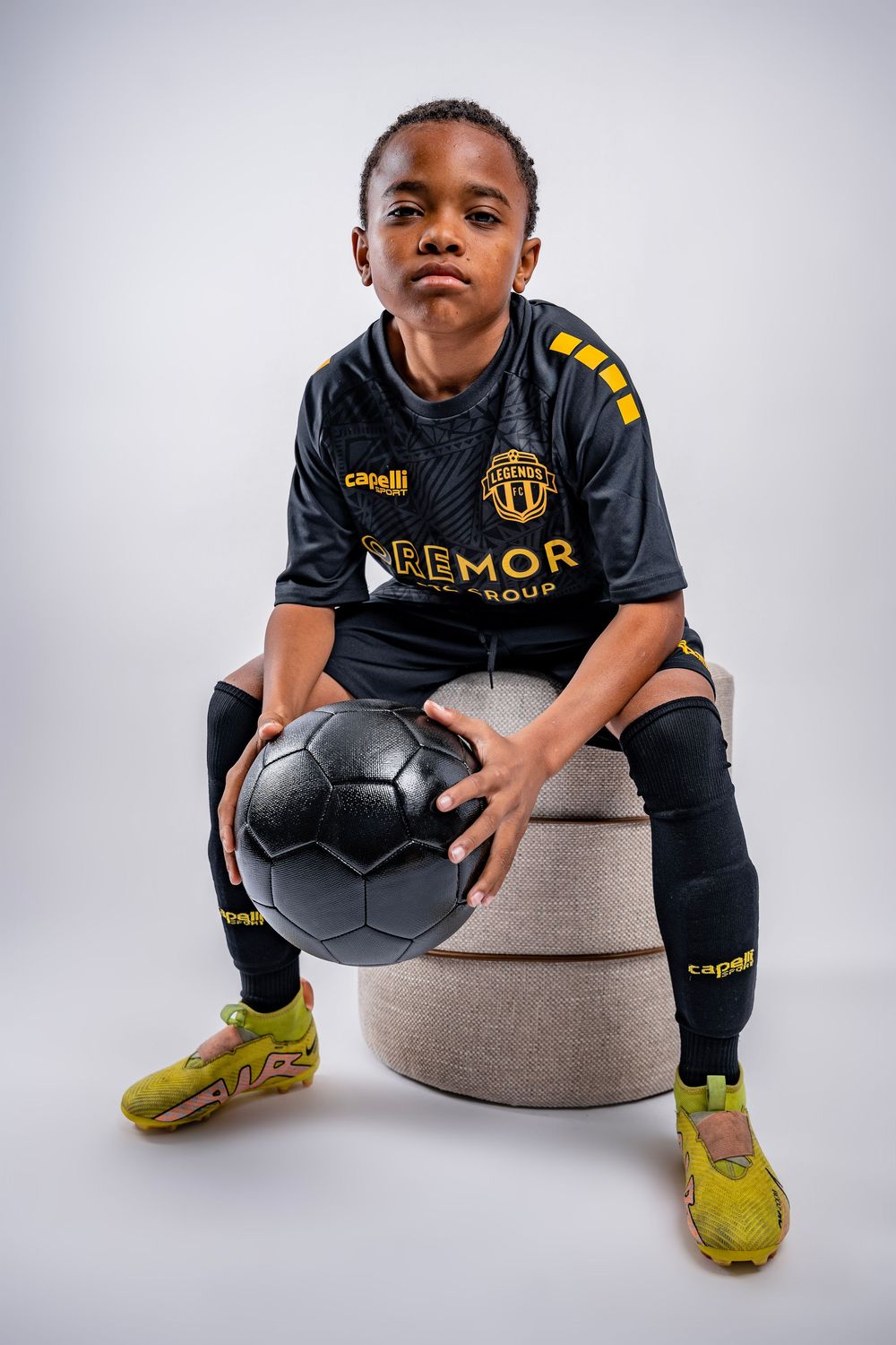 A young athlete in a black Aemor jersey and yellow shoes poses with a soccer ball on a gray ottoman in studio lighting.