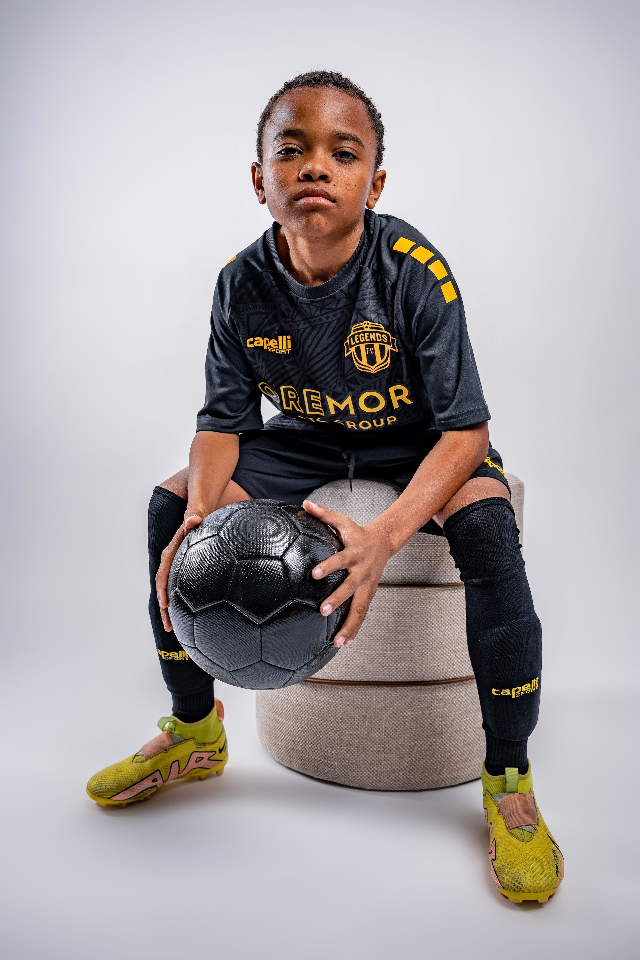 A young athlete in a black Aemor jersey and yellow shoes poses with a soccer ball on a gray ottoman in studio lighting.