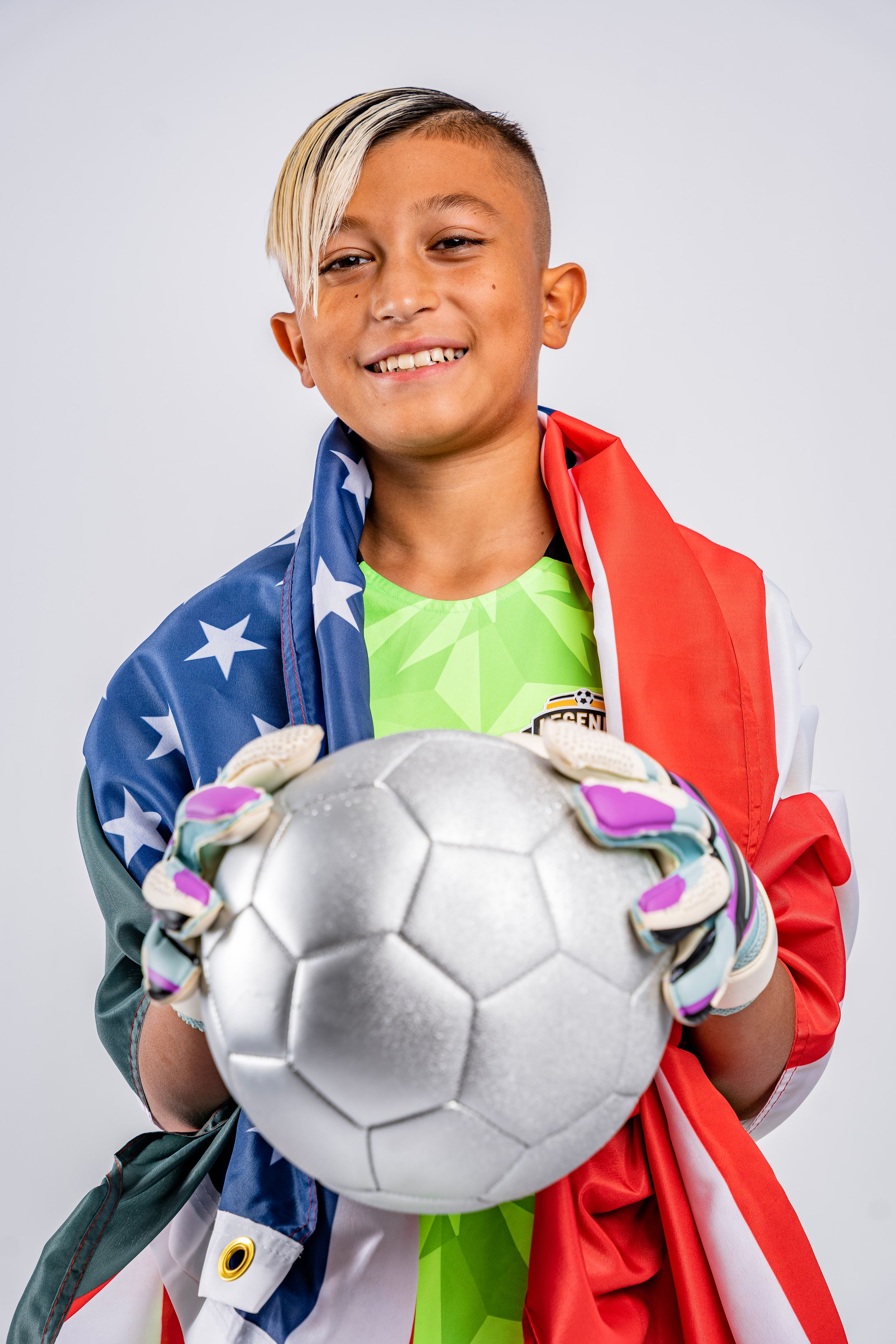 Young athlete wrapped in American flag smiles while holding silver soccer ball against white background.