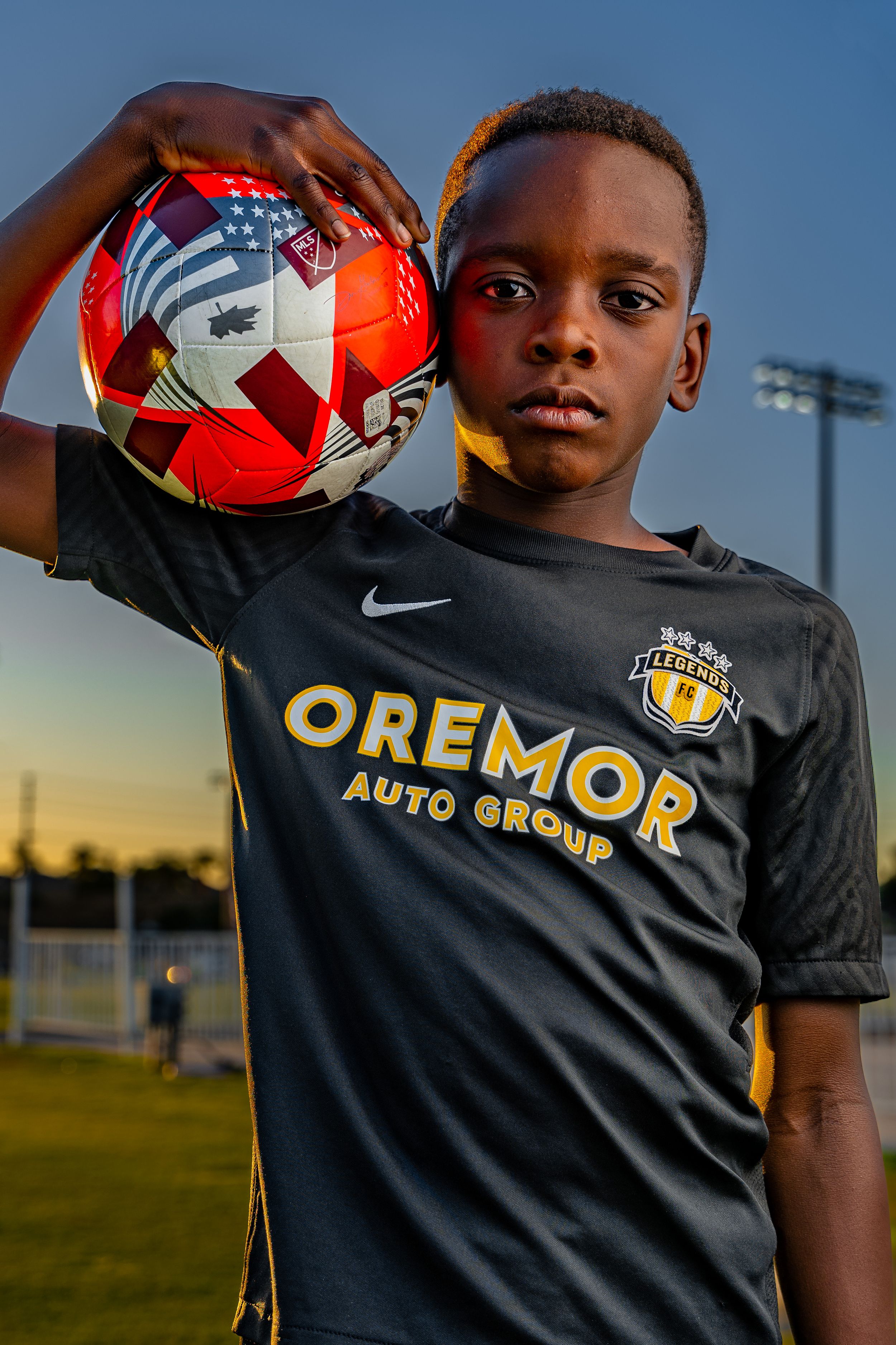 Soccer player wearing black Oremor Auto Group jersey balancing ball on shoulders during sunset practice session.