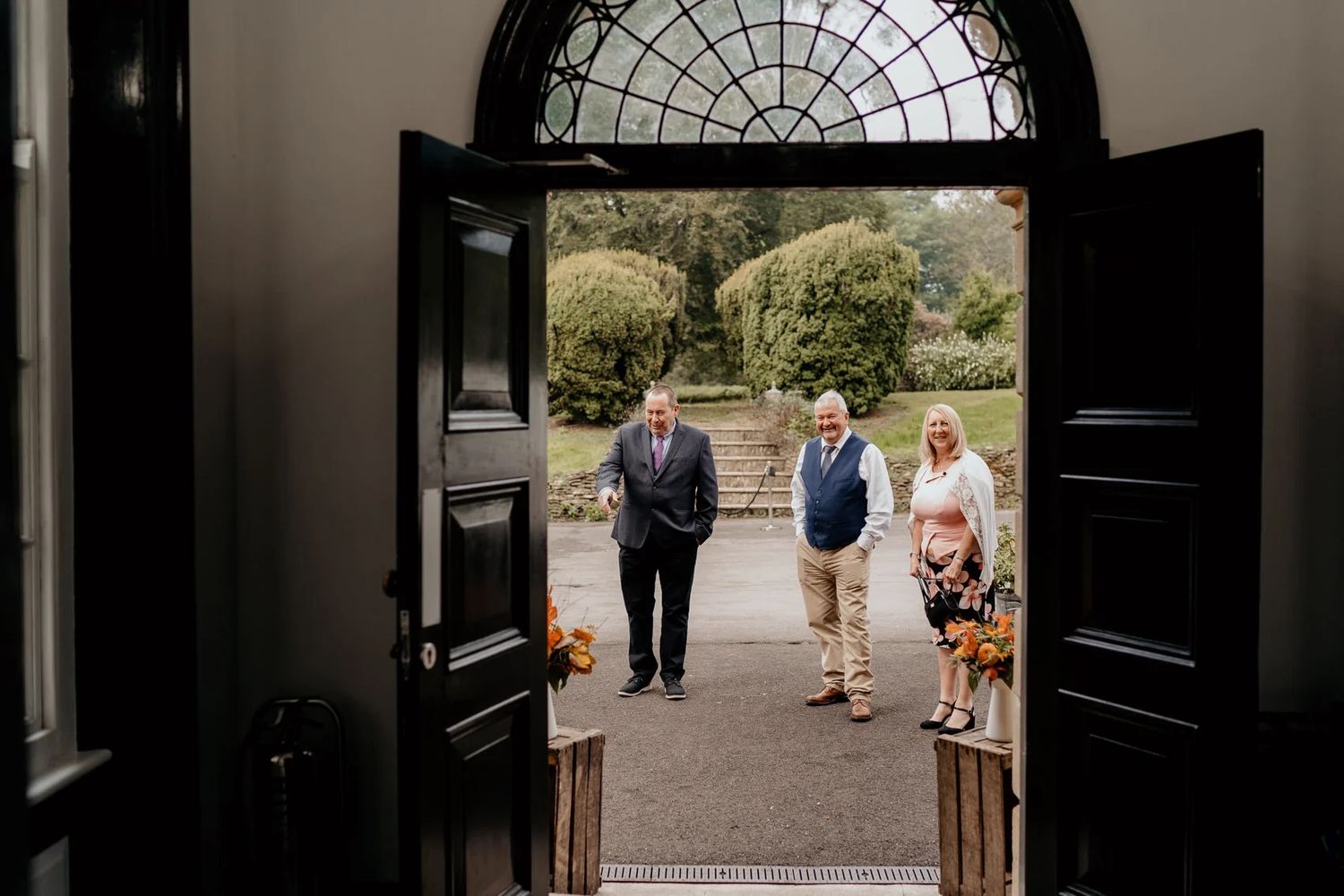 Through grand open black double doors, a group stands with their dog on a garden path beneath an arched window.