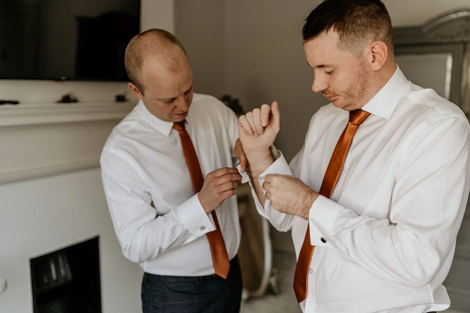 Groomsmen in white shirts and orange ties help each other get ready for a wedding celebration.