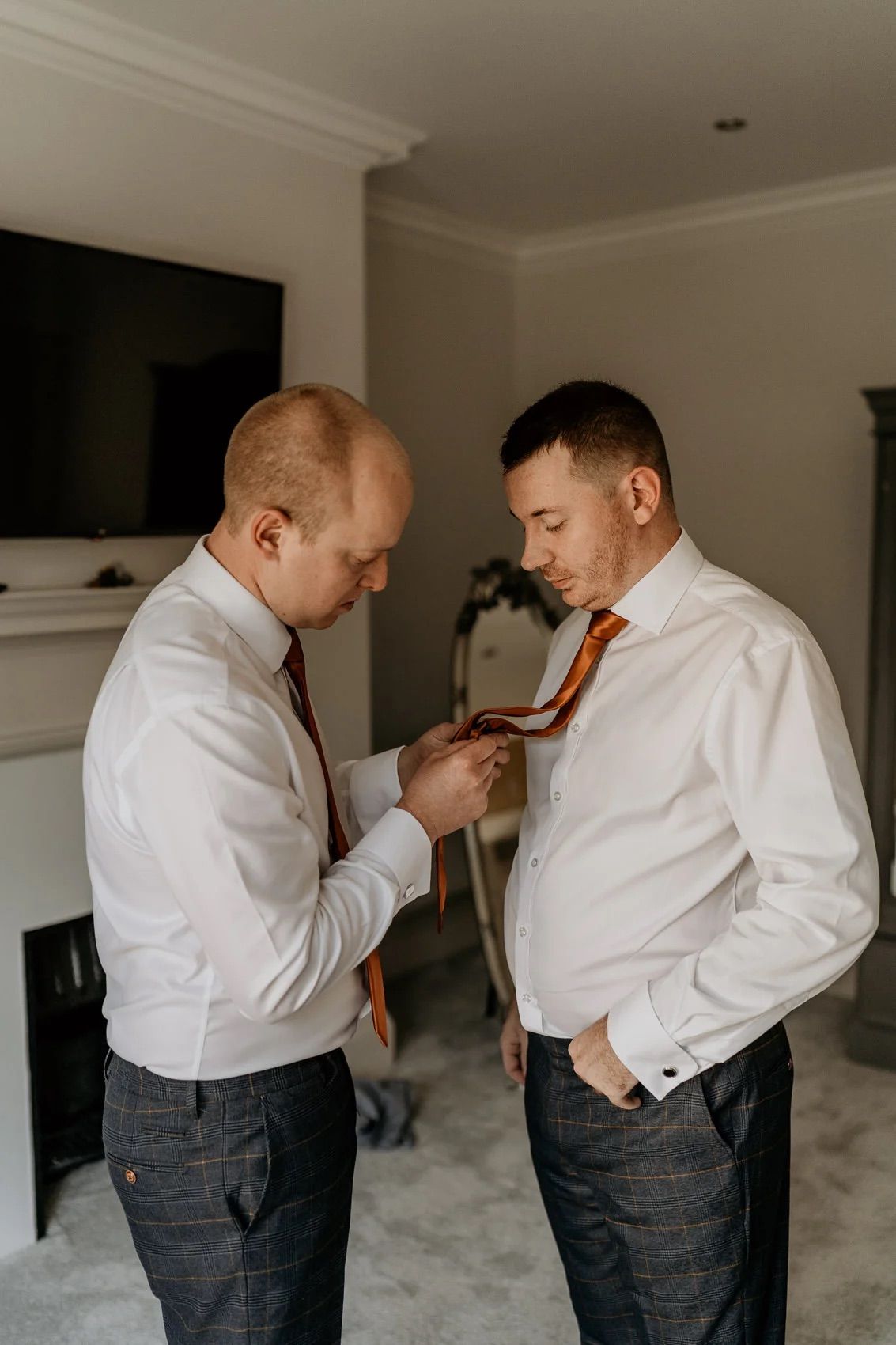 Two people in white dress shirts and gray pants help each other adjust their neckties in a stylish interior setting.