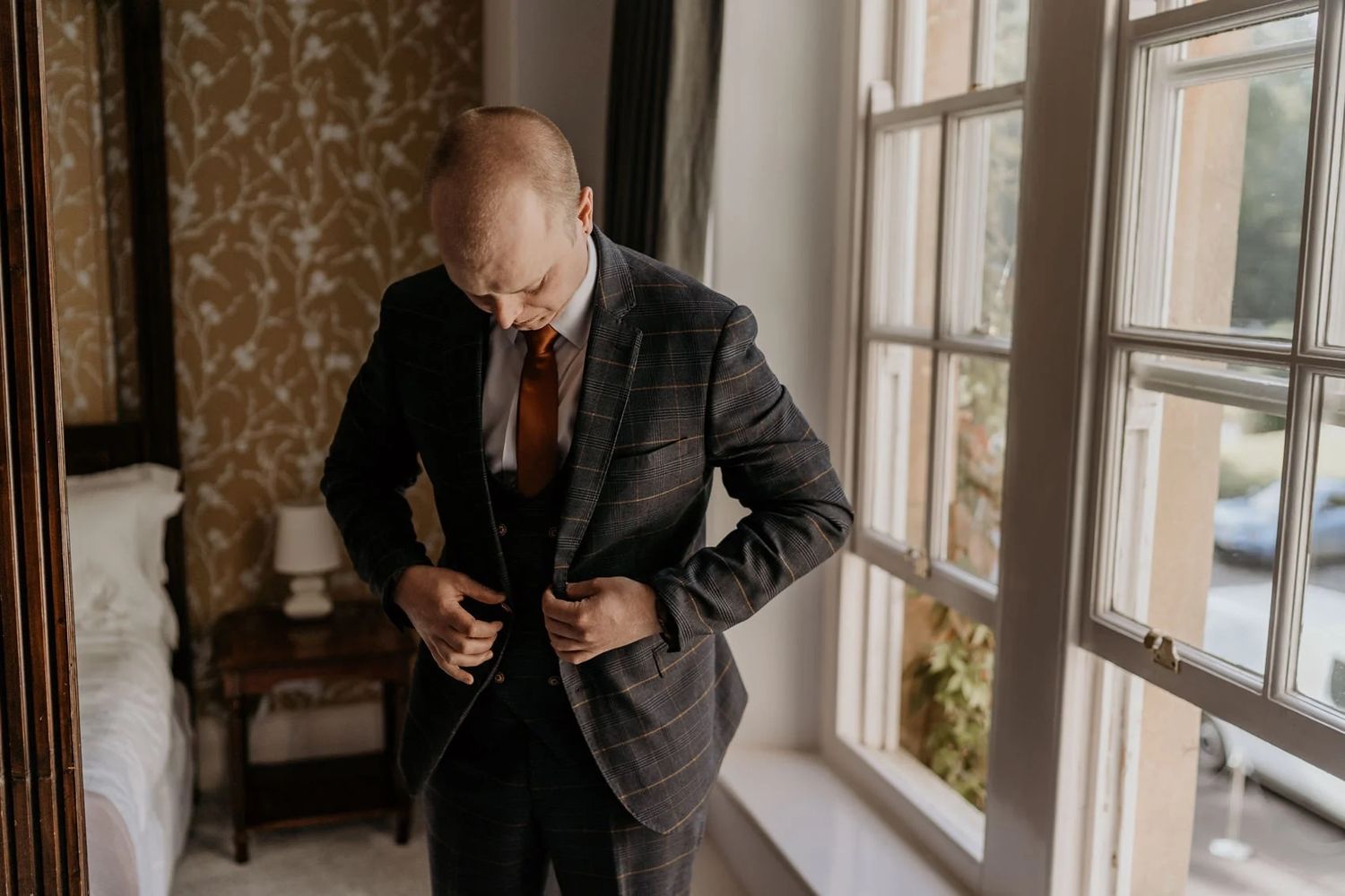 A person in a formal suit adjusts their tie while standing near sunlit windows in an elegant room.