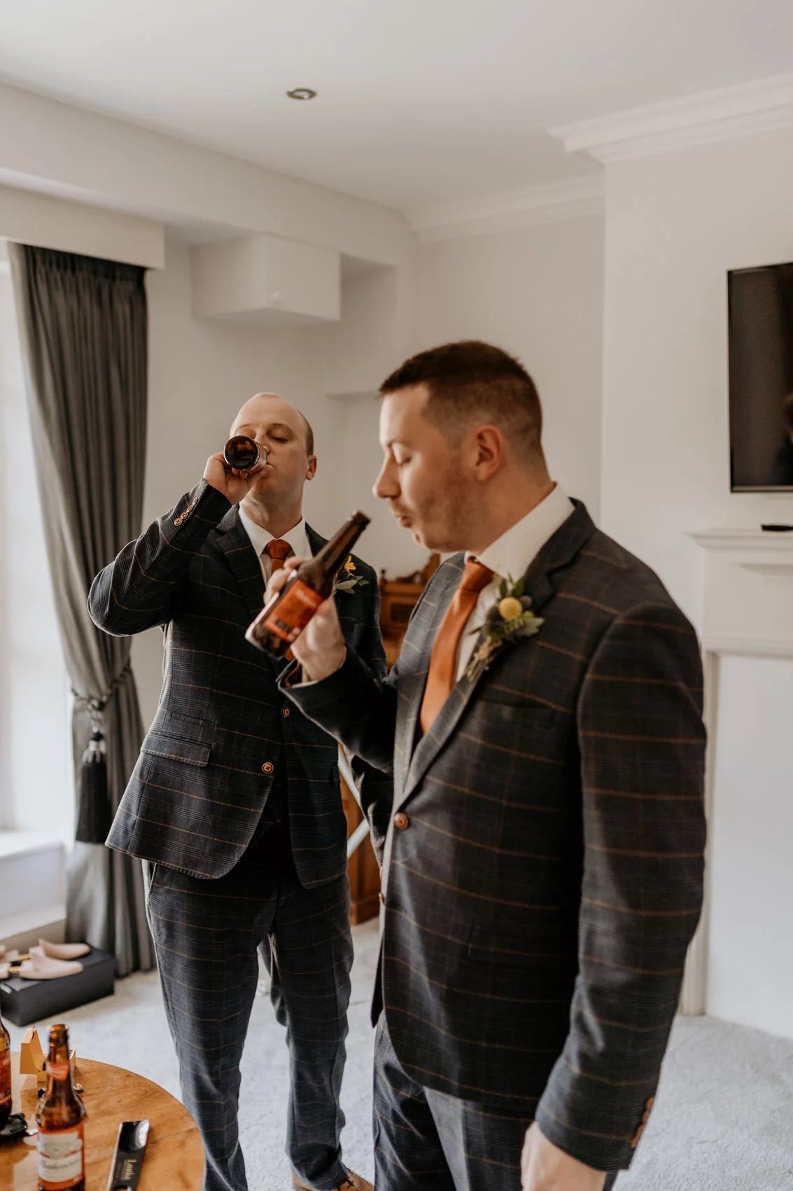 Groomsmen in plaid suits sharing drinks during pre-wedding preparations in a bright hotel room.