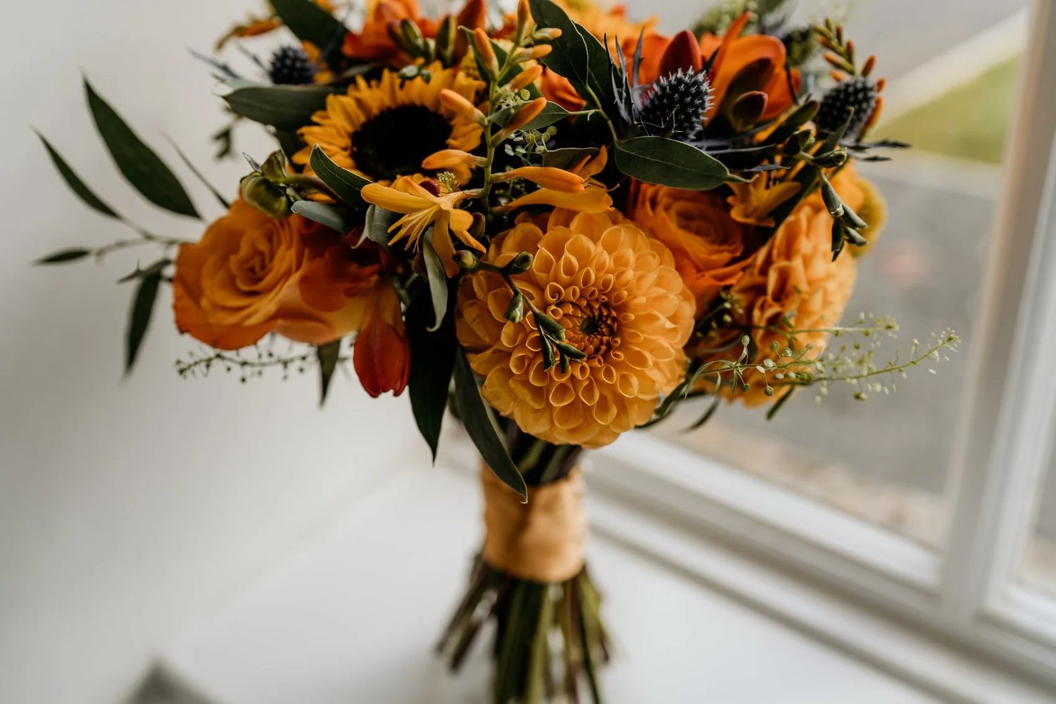 Vibrant orange and yellow sunflower wedding bouquet with roses and greenery sits by a bright window.