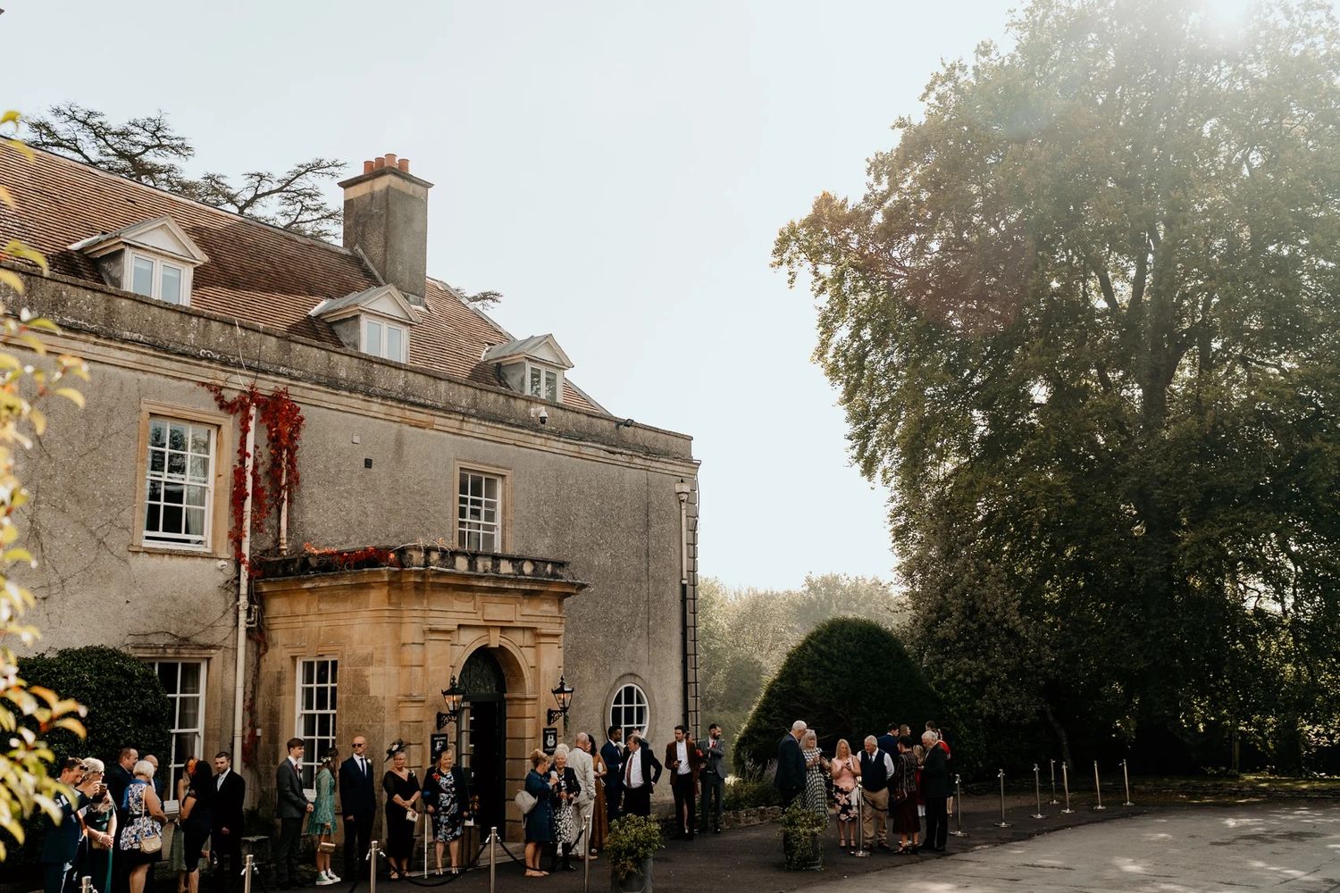 Panoramic exterior view of a historic mansion wedding venue with guests gathered outside during golden hour.