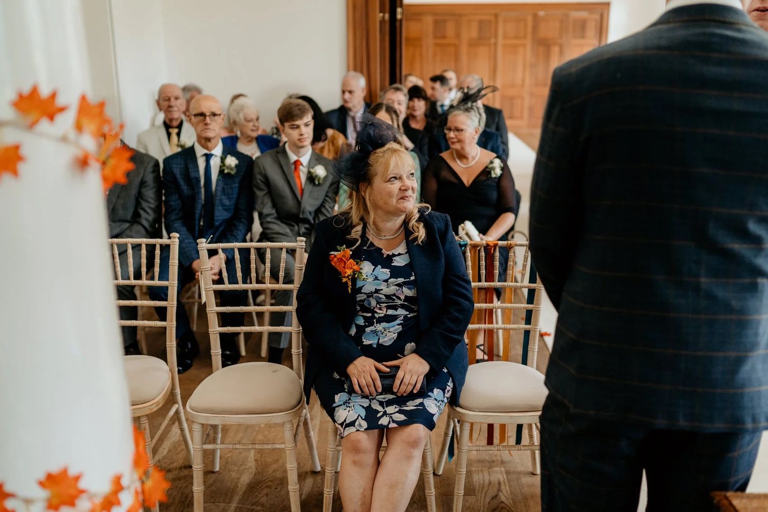 Wedding guests seated in white Chiavari chairs under a crystal chandelier during an indoor ceremony.