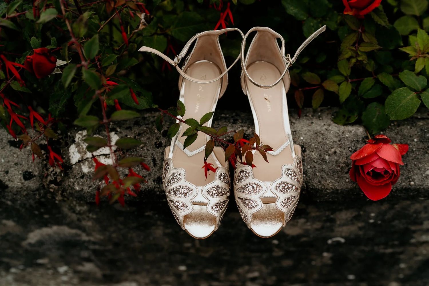 Elegant white bridal heels arranged in a row beside blooming red roses against a stone backdrop.