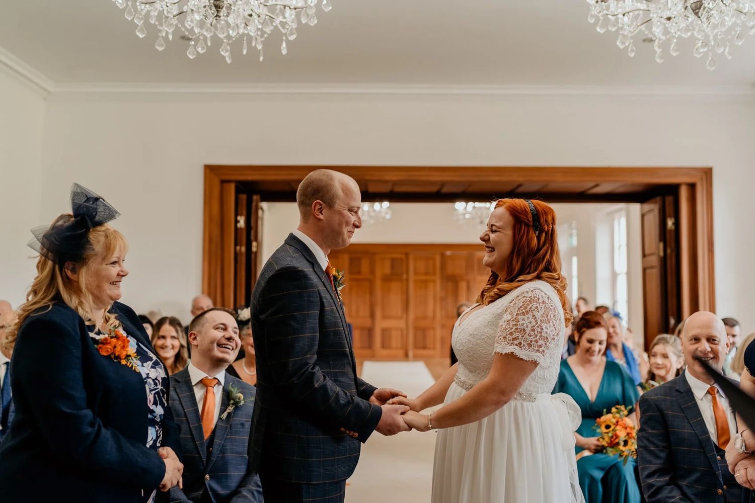 Wedding ceremony taking place in an elegant white room with crystal chandeliers overhead and guests seated in rows.