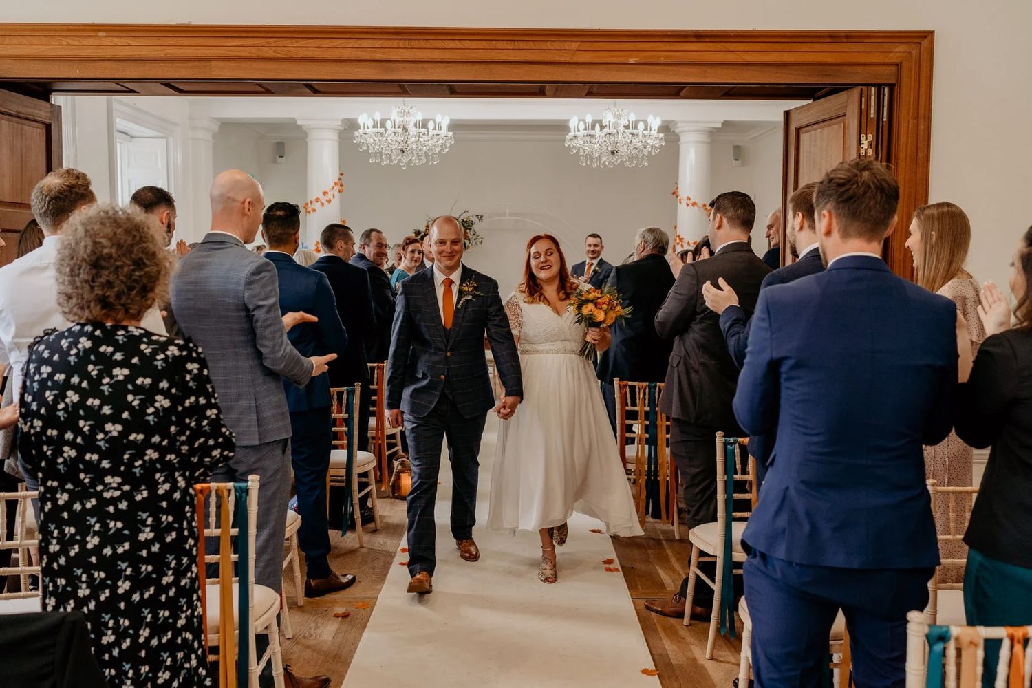 Wedding party processes down aisle in bright indoor venue with chandelier overhead.