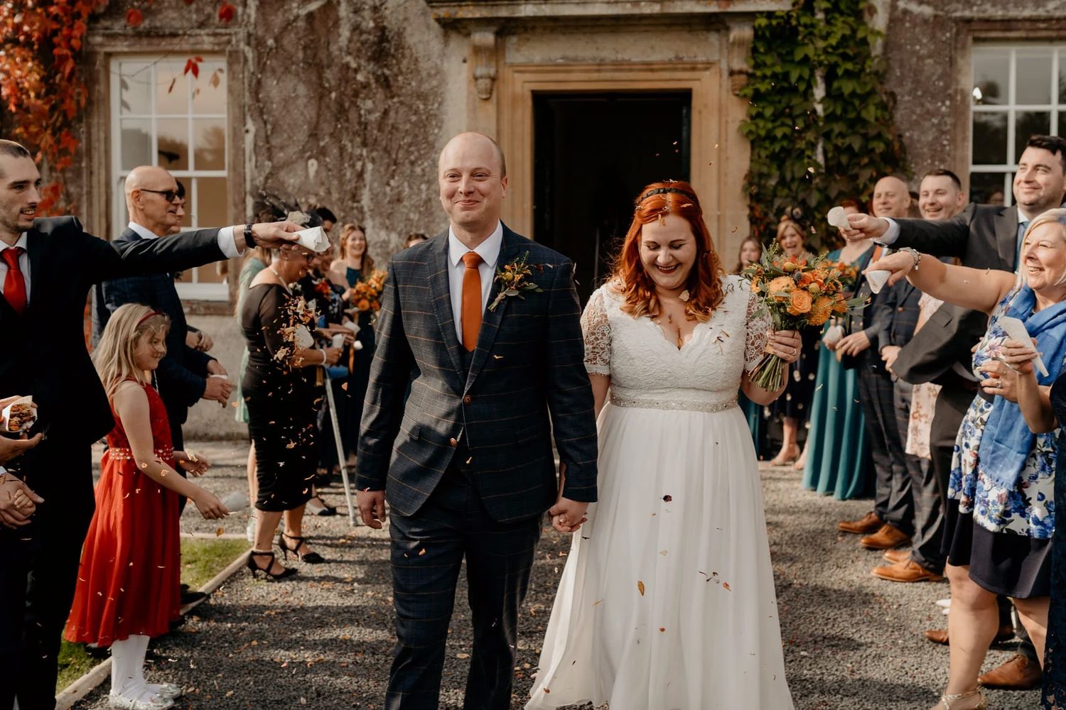 Wedding ceremony guests gather outdoors on a stone walkway surrounded by autumn foliage at a historic venue.