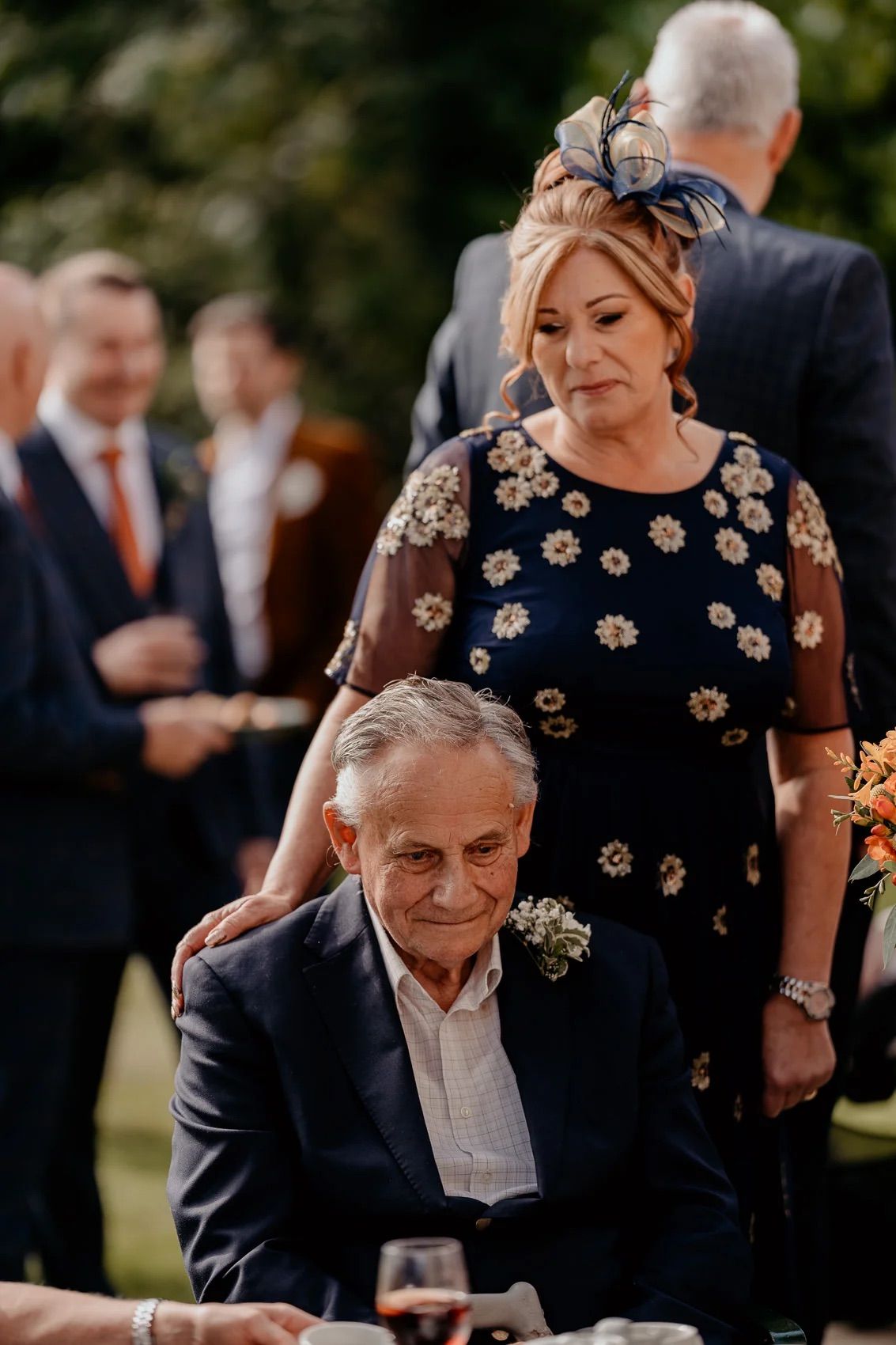An outdoor wedding scene shows guests in formal attire gathered on a sunny autumn day.