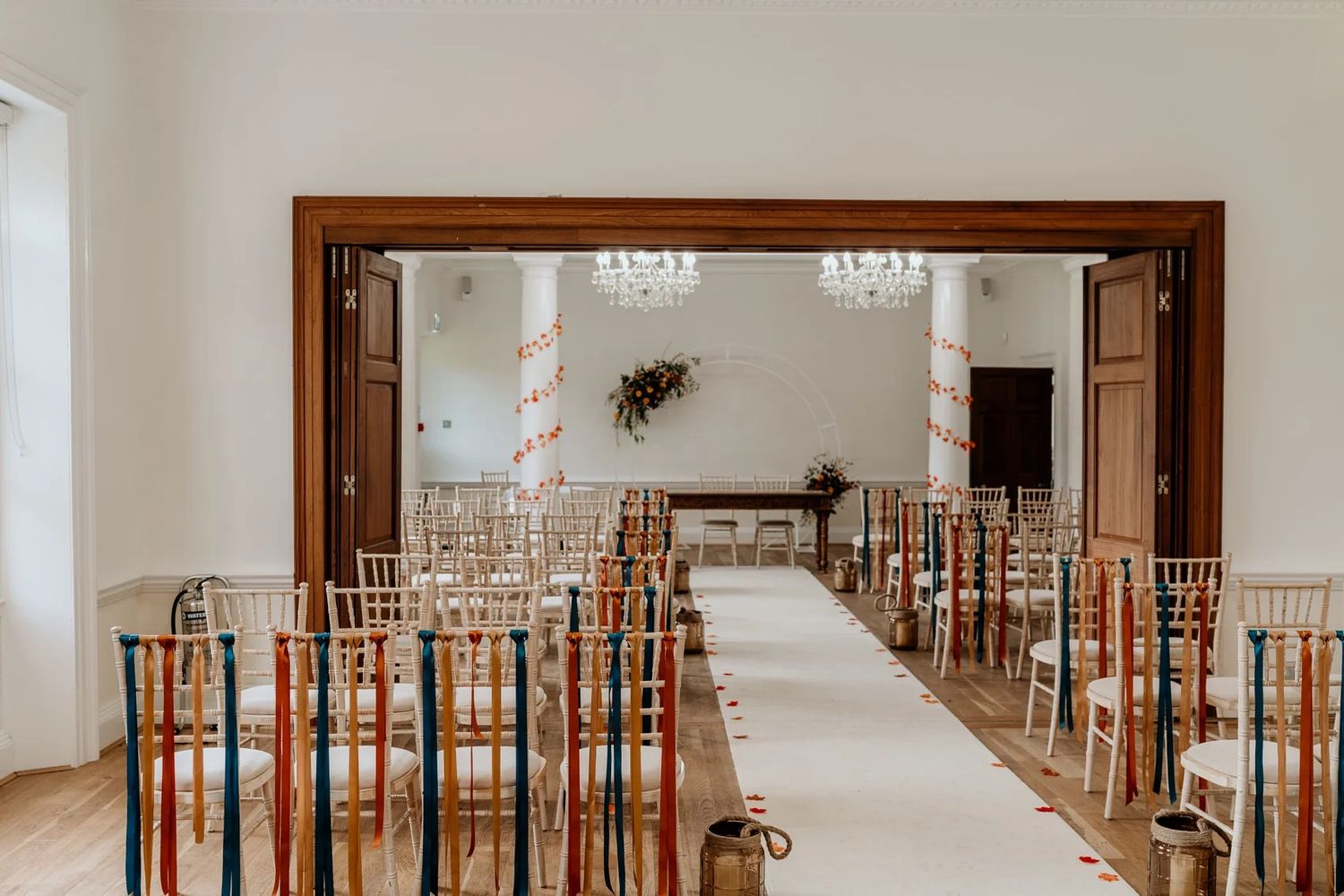 A long empty wedding aisle with white walls and wooden chairs lined up leading to a bright ceremony space.