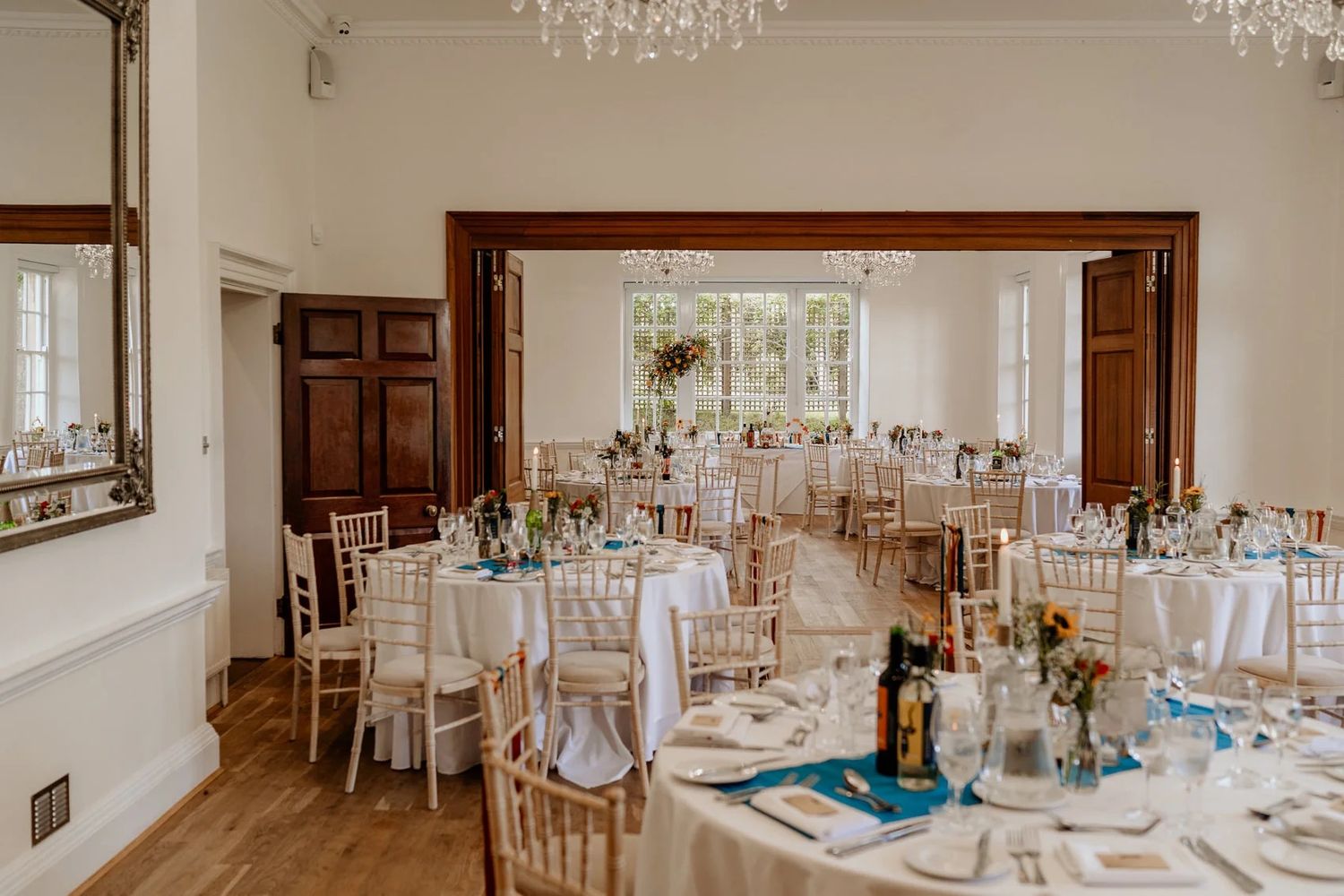 Elegant wedding reception hall with crystal chandeliers and white tables set for dining in a bright interior space.