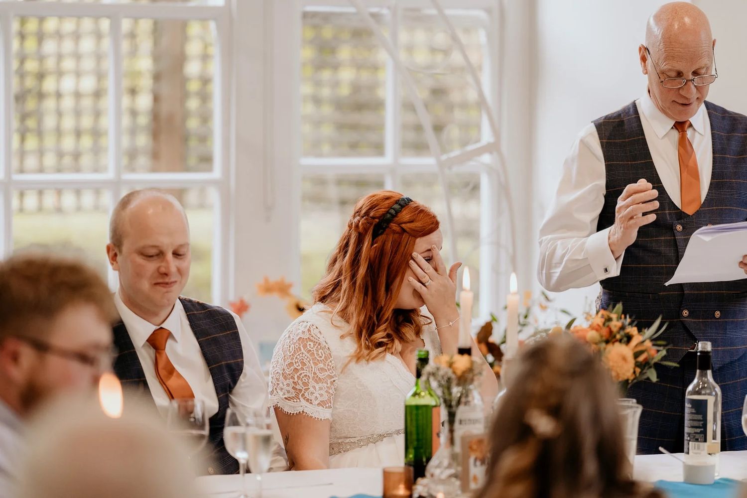 Sunlit wedding reception with guests seated at long tables decorated with floral centerpieces.