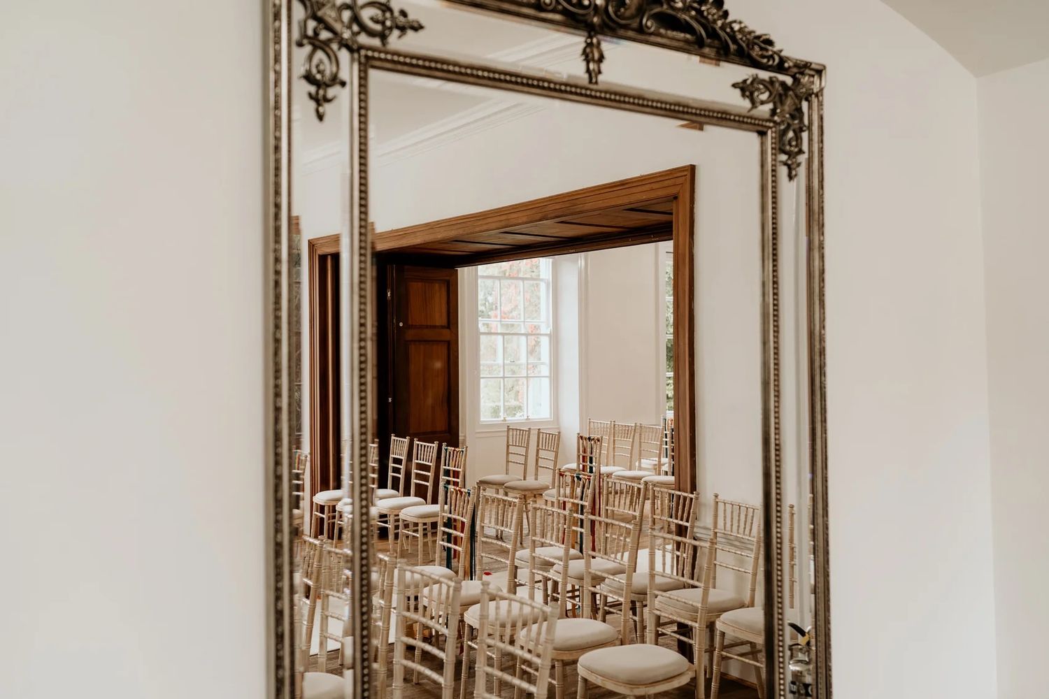 Ornate gold mirror hangs on white wall next to empty dining room with natural light streaming through windows.