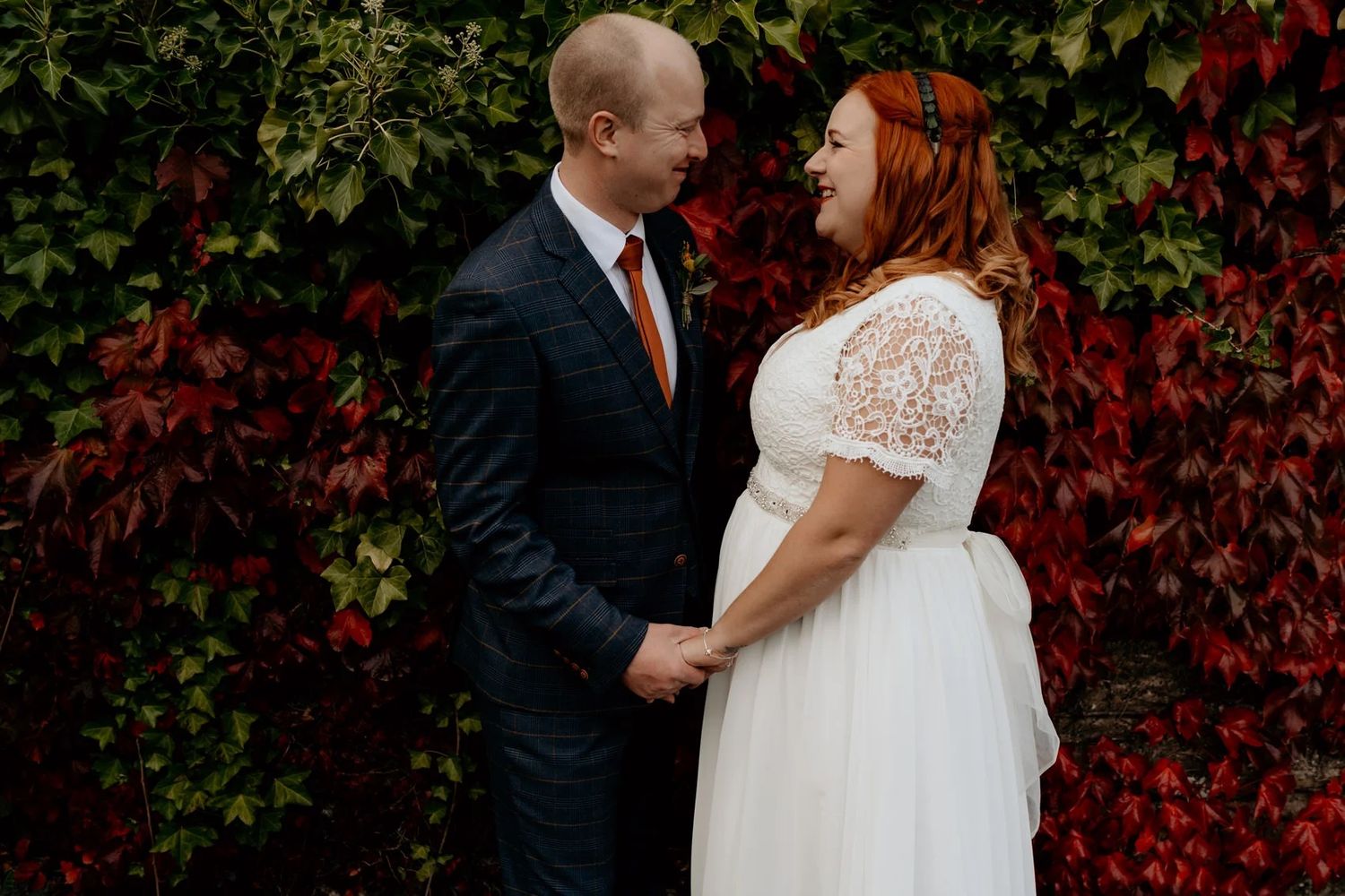Newlyweds embrace against a backdrop of vibrant red autumn foliage near a brick building.