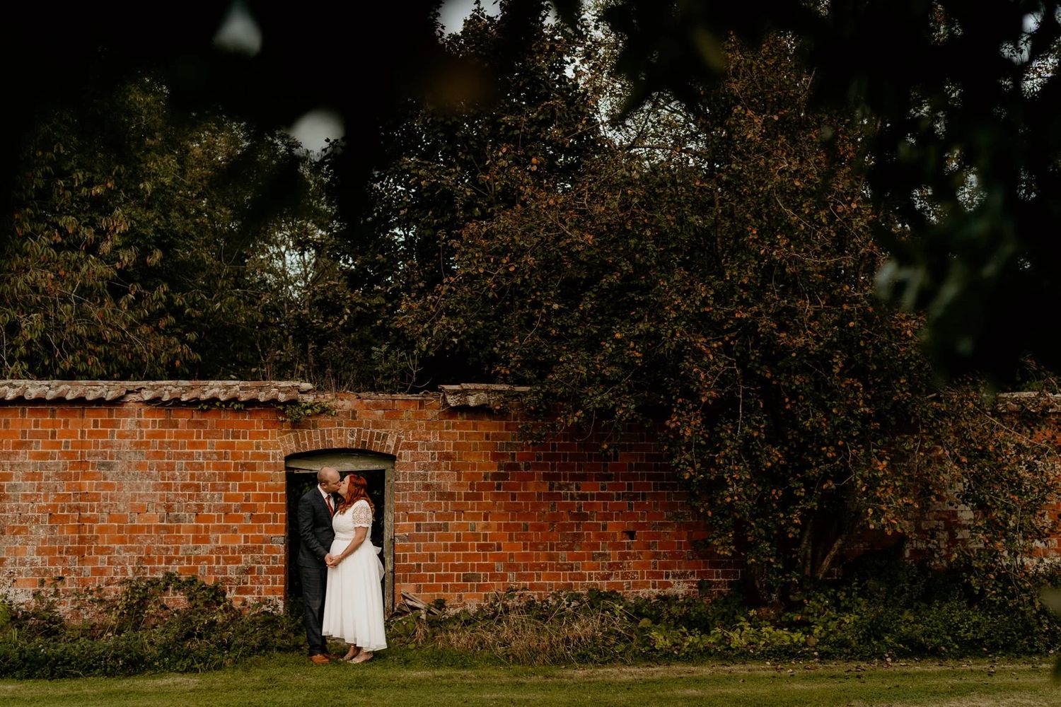 A couple framed by a rustic red brick garden wall archway with lush greenery overhead.