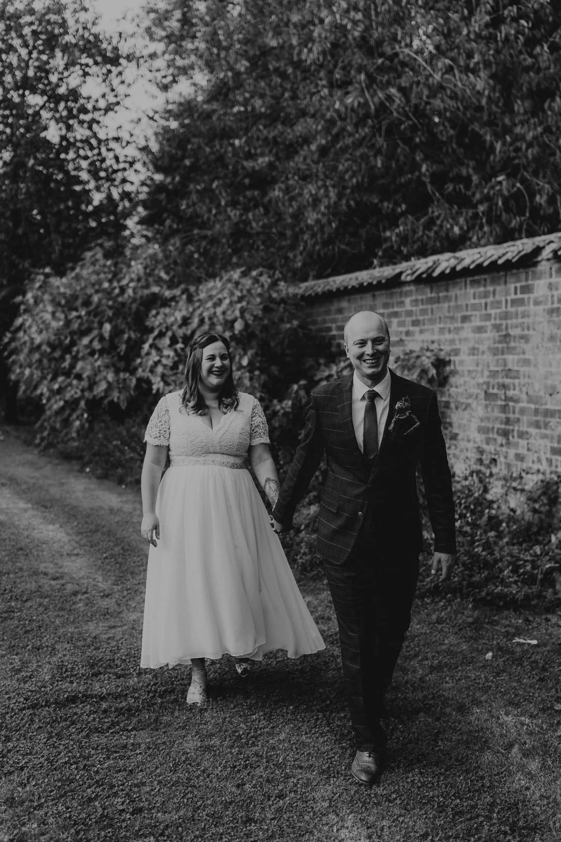 A couple walks together on a path alongside a brick wall and lush greenery in black and white.