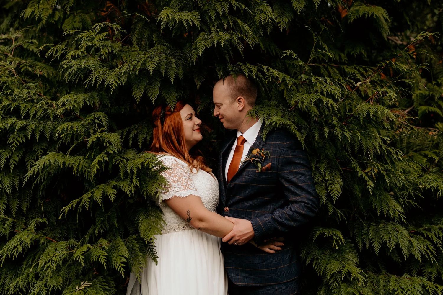 A couple shares an intimate moment amid lush evergreen trees during their wedding celebration.
