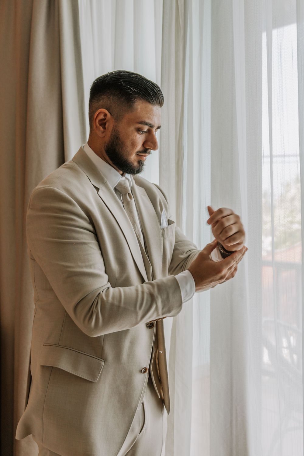 A groom in a beige suit adjusts his cuff while standing near a window with sheer curtains.