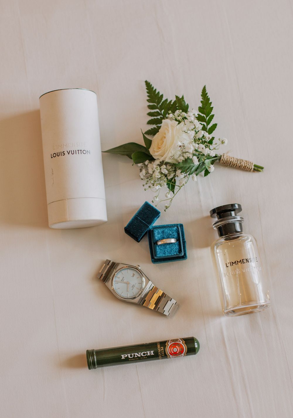 Wedding accessories including perfume bottles, Louis Vuitton packaging, delicate flowers, and a wristwatch on white surface.
