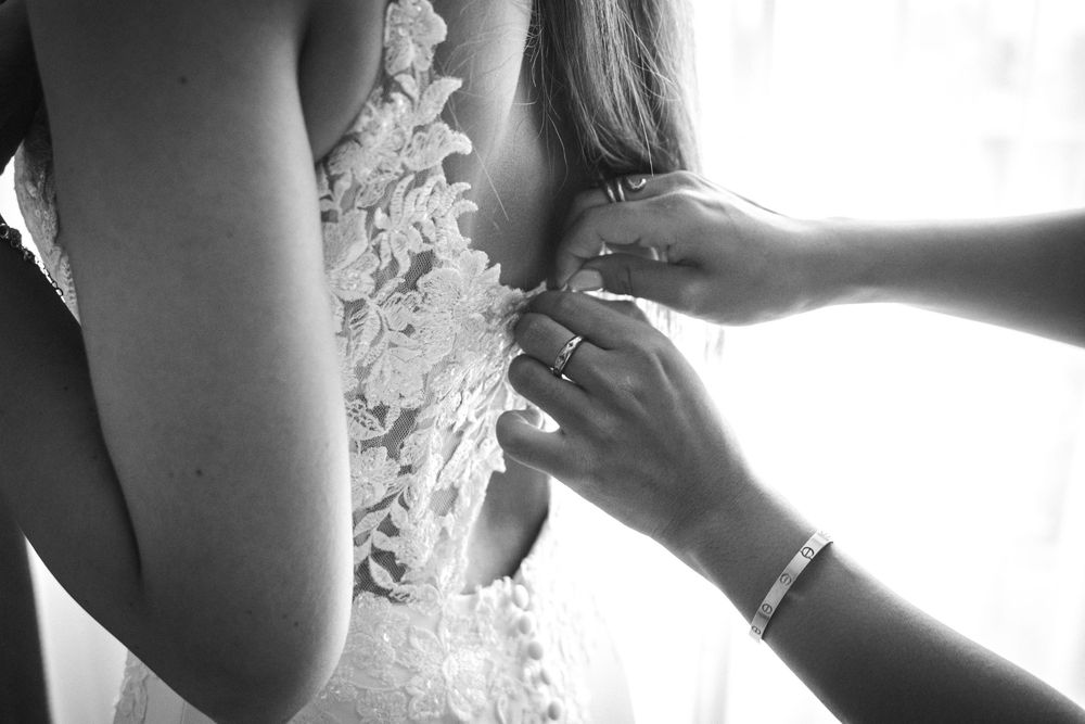 Close-up black and white photo showing hands buttoning delicate lace details on a wedding dress.